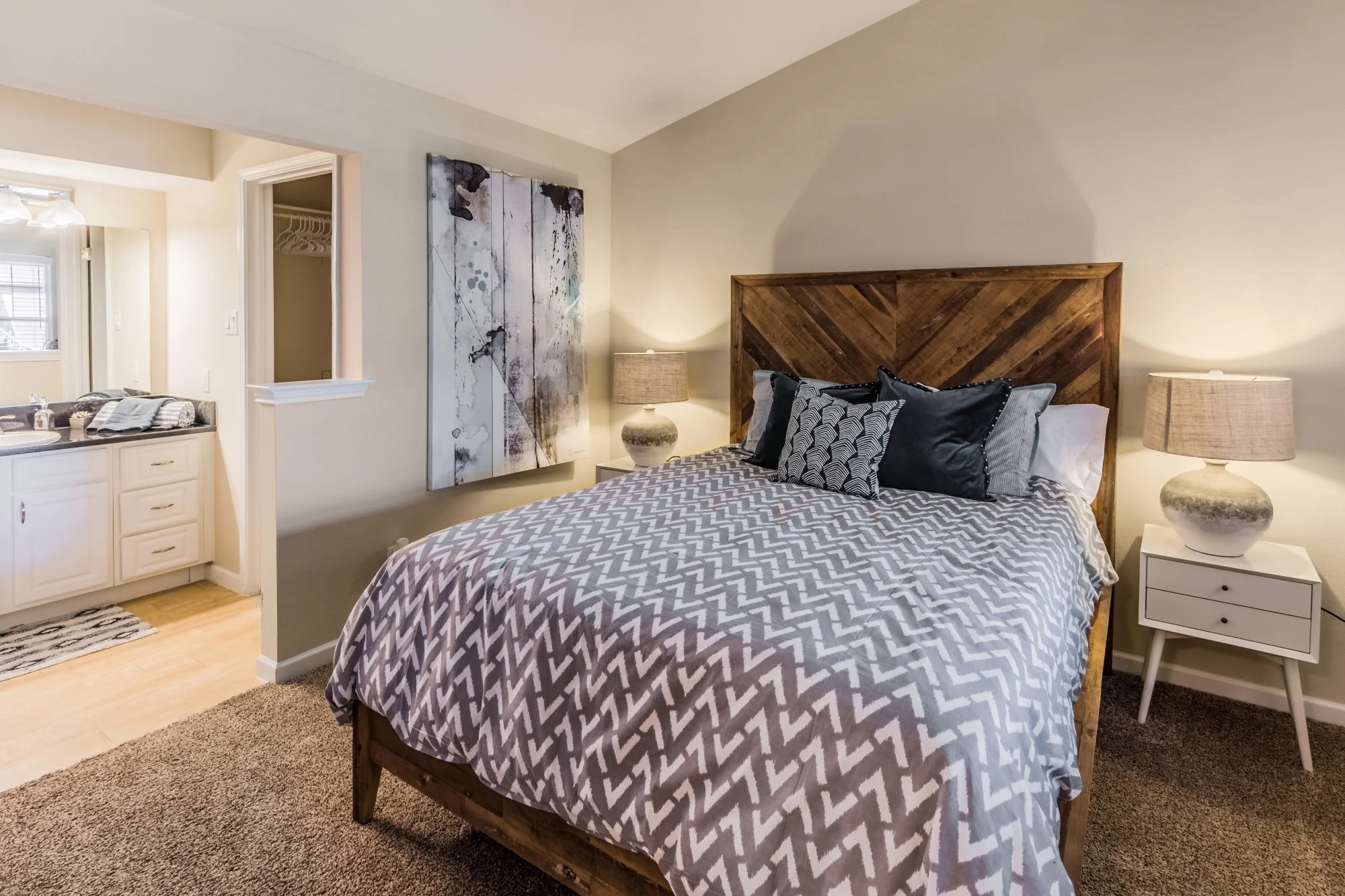 A cozy bedroom with a wooden bed frame, patterned bedding, and pillows. There are two matching bedside lamps and a large abstract art piece on the wall. The room has beige walls and carpeted floor. Part of a bathroom with white cabinetry and a window