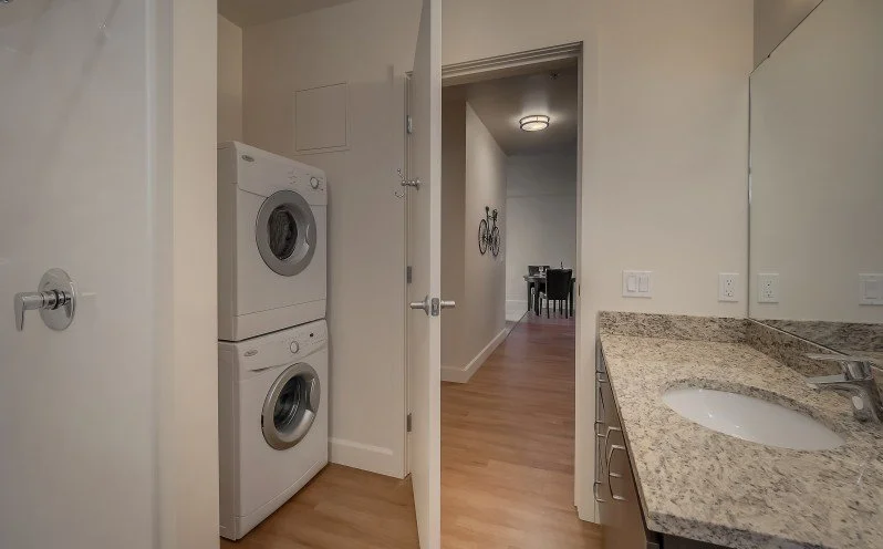 A laundry area with a stacked washer and dryer beside a door, next to a bathroom vanity with a granite countertop and sink, with hallway and dining area in the background.