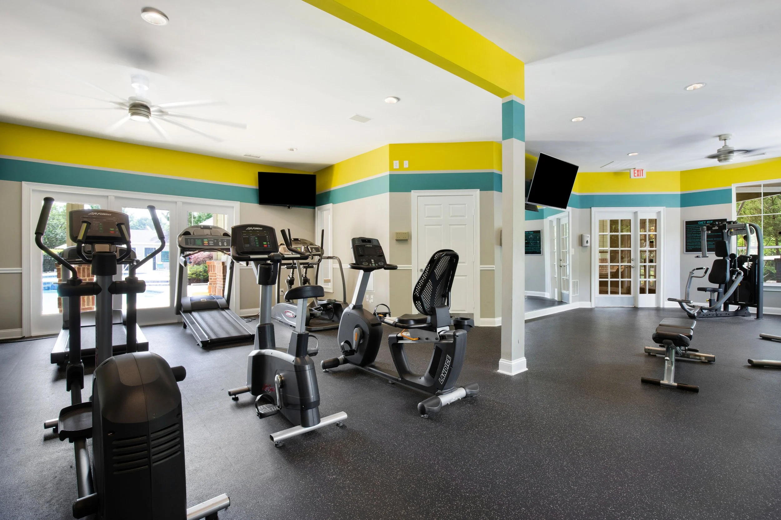 Indoor gym with cardio equipment including treadmills and stationary bikes, bright walls with colorful stripes, ceiling fans, and large windows.