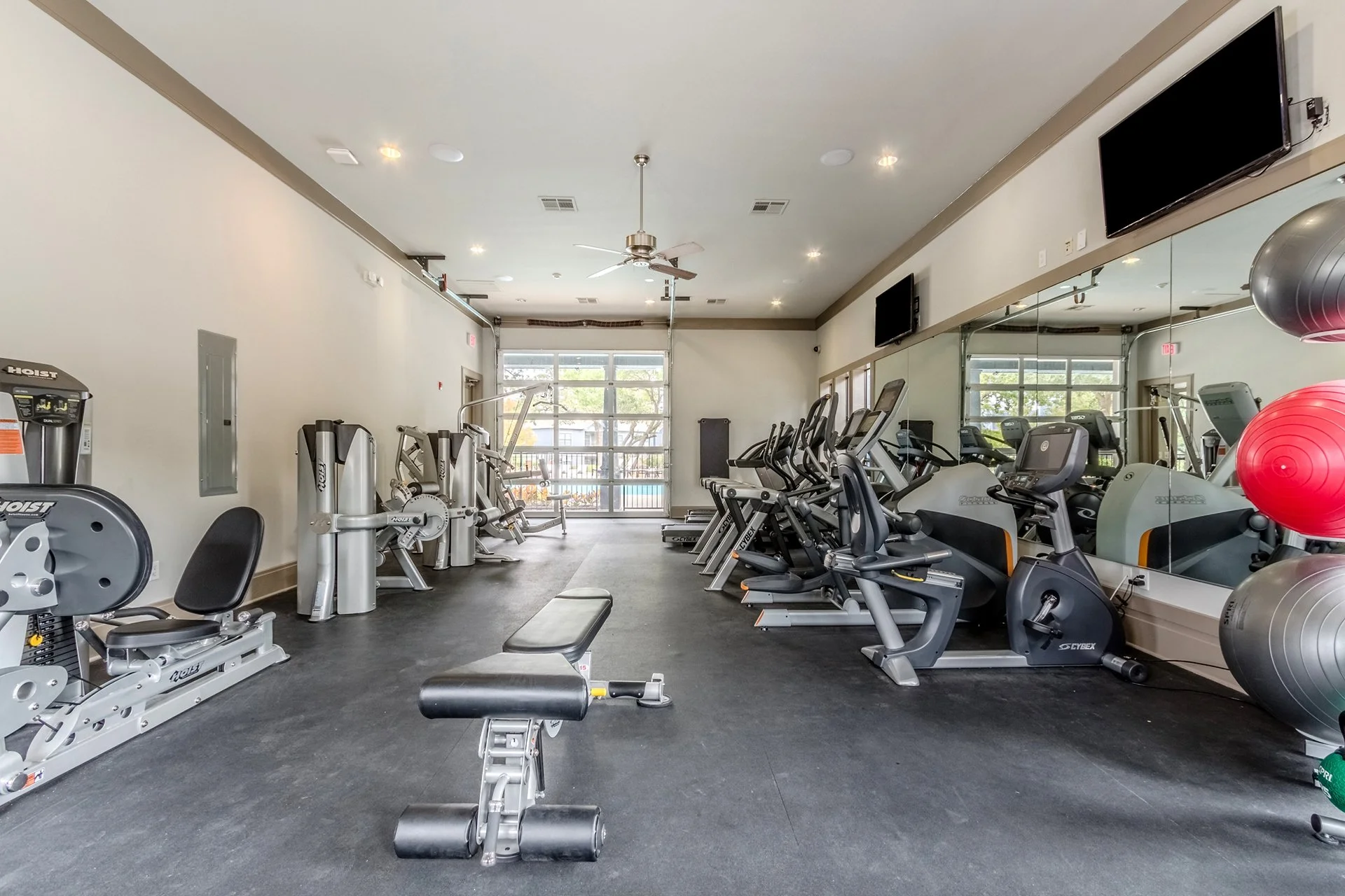 Empty gym with various exercise equipment including treadmills, stationary bikes, weight machines, and stability balls, with large windows and a mirror wall.