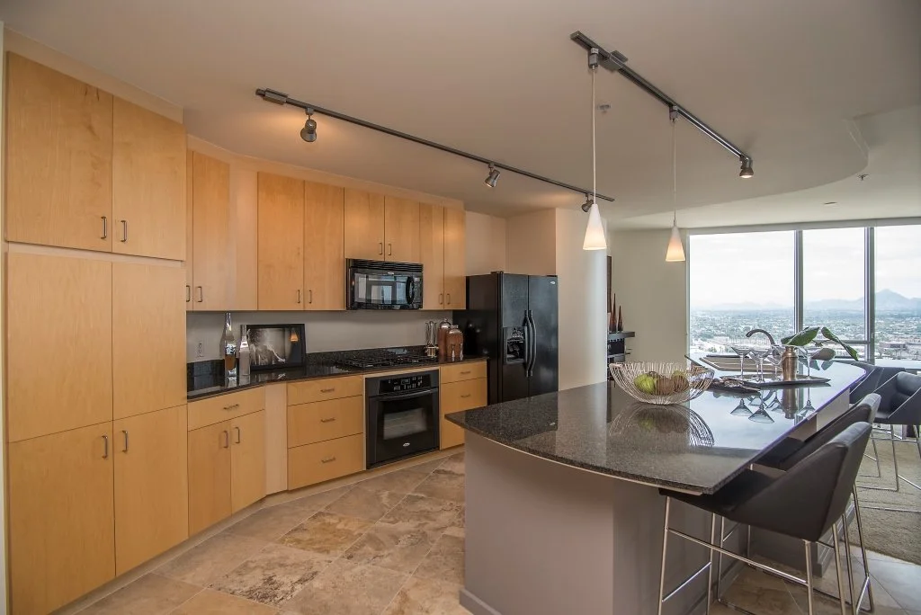 Modern kitchen with light wood cabinets, black appliances, a black refrigerator, and a curved granite countertop island with bar stools. Large windows offer a city view.