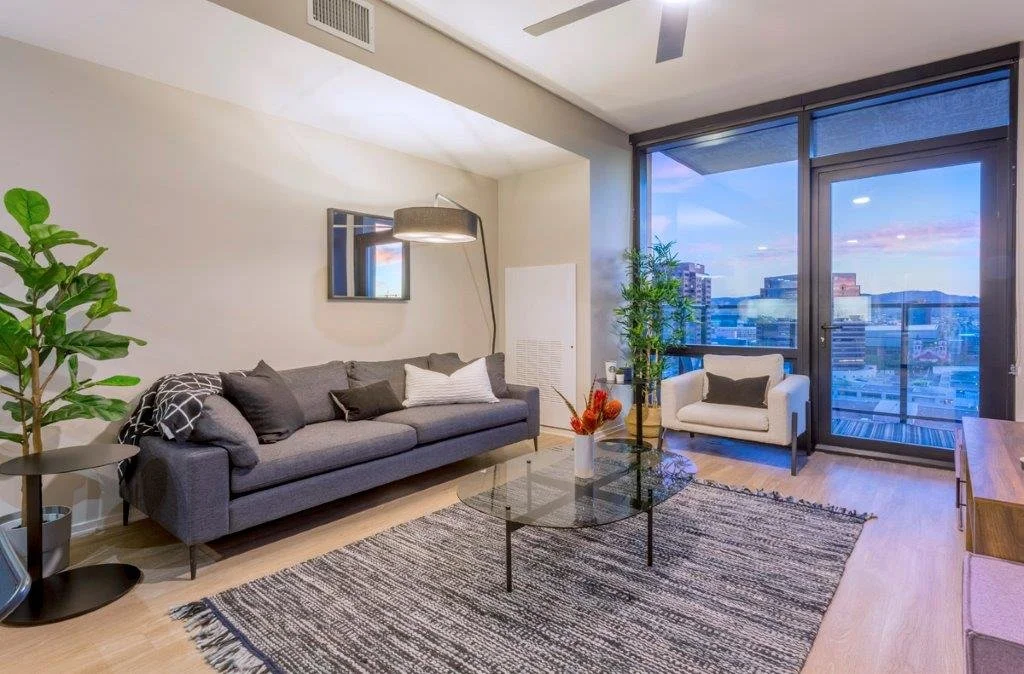 Modern living room with a gray sofa, white armchair, glass coffee table, indoor plants, and large windows showing city buildings and a view of the city skyline at sunset.