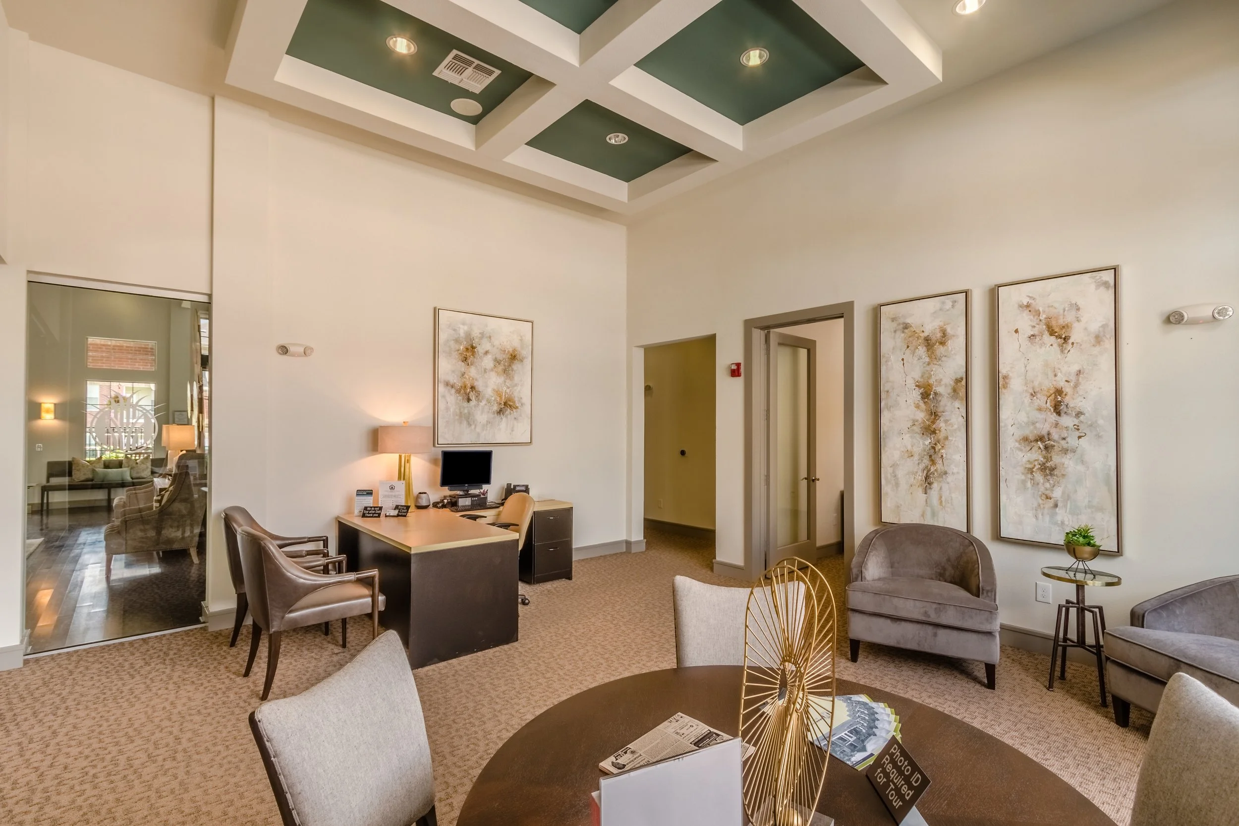 A spacious modern waiting or lounge area with a round table, chairs, a desk with a computer, chairs along the wall, abstract paintings on the wall, and decorative lighting on the ceiling.