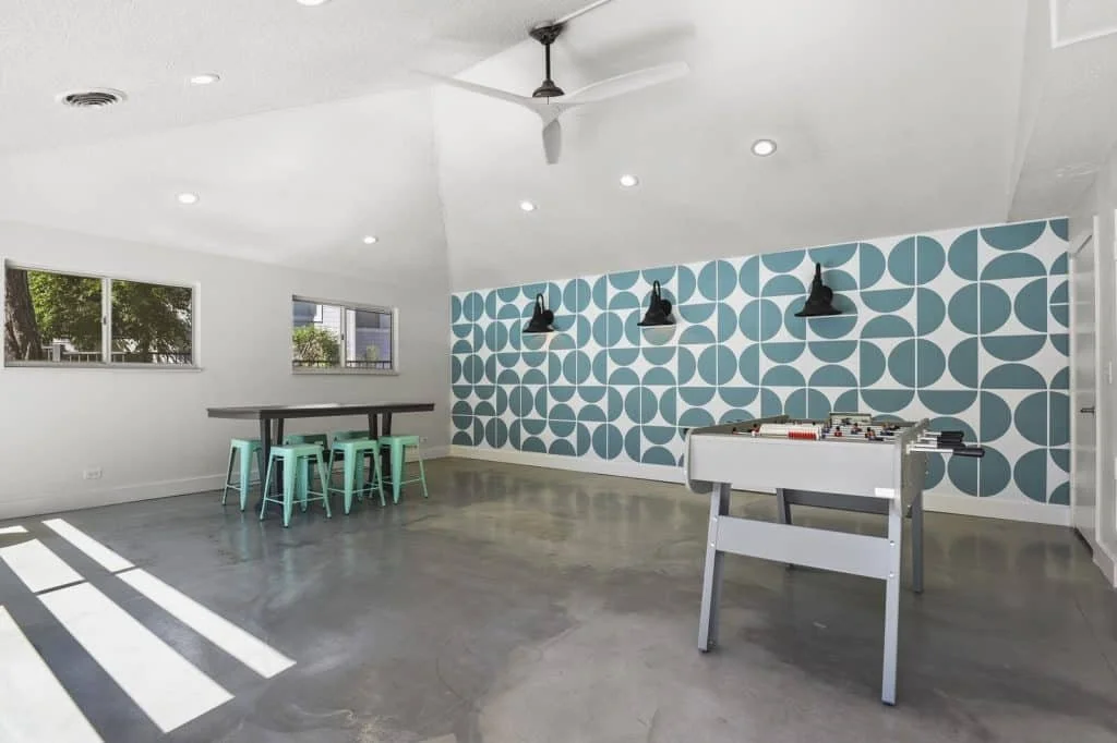 A modern, minimalistic indoor recreational space with a foosball table, a long table with mint green stools, windows showing trees outside, a decorative blue geometric wall, black pendant lights, ceiling fan, and concrete flooring.