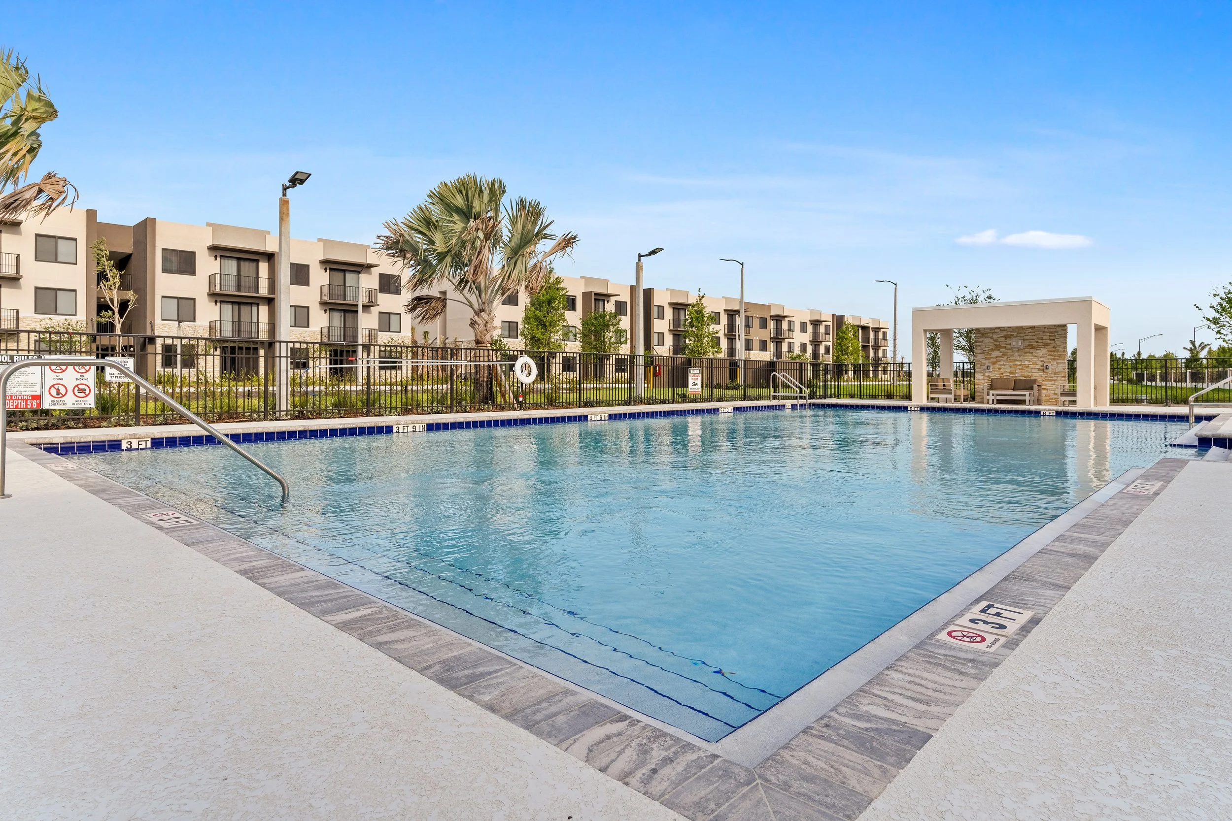 Empty outdoor swimming pool with a surrounding concrete deck, black fence, apartment buildings in the background, and a small covered seating area with benches, under a clear blue sky.