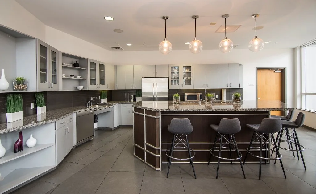 Modern kitchen with white and gray cabinets, stainless steel appliances, granite countertops, and a central island with four gray barstools. Pendant lights hang above the island, and decorative items like vases and potted plants are displayed on coun