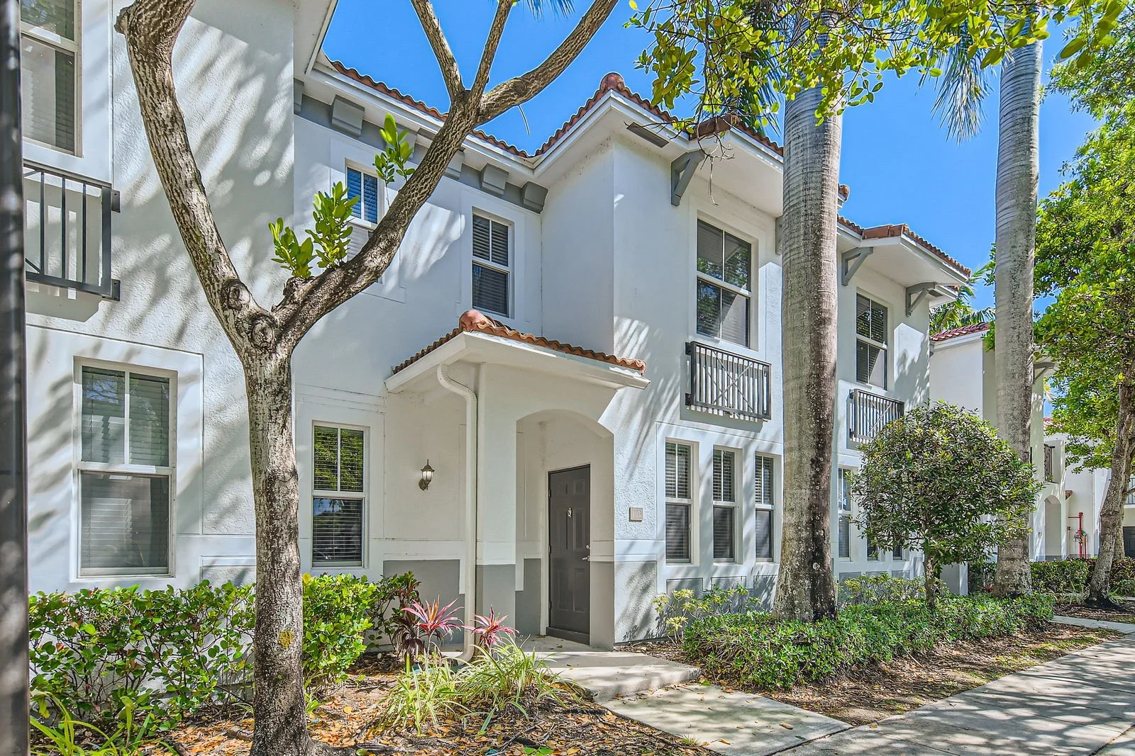 White residential building surrounded by trees and plants, with multiple windows, small balconies, and a sidewalk in front.