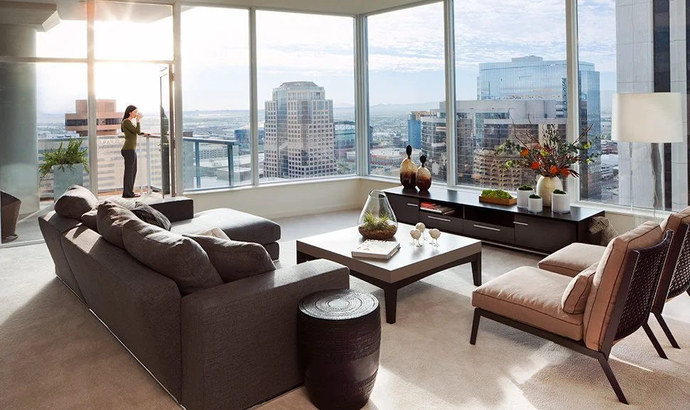 Modern high-rise apartment living room with large windows overlooking city skyline, featuring gray sofa, pink armchair, coffee table with decorative items, and a woman on balcony taking a photo.