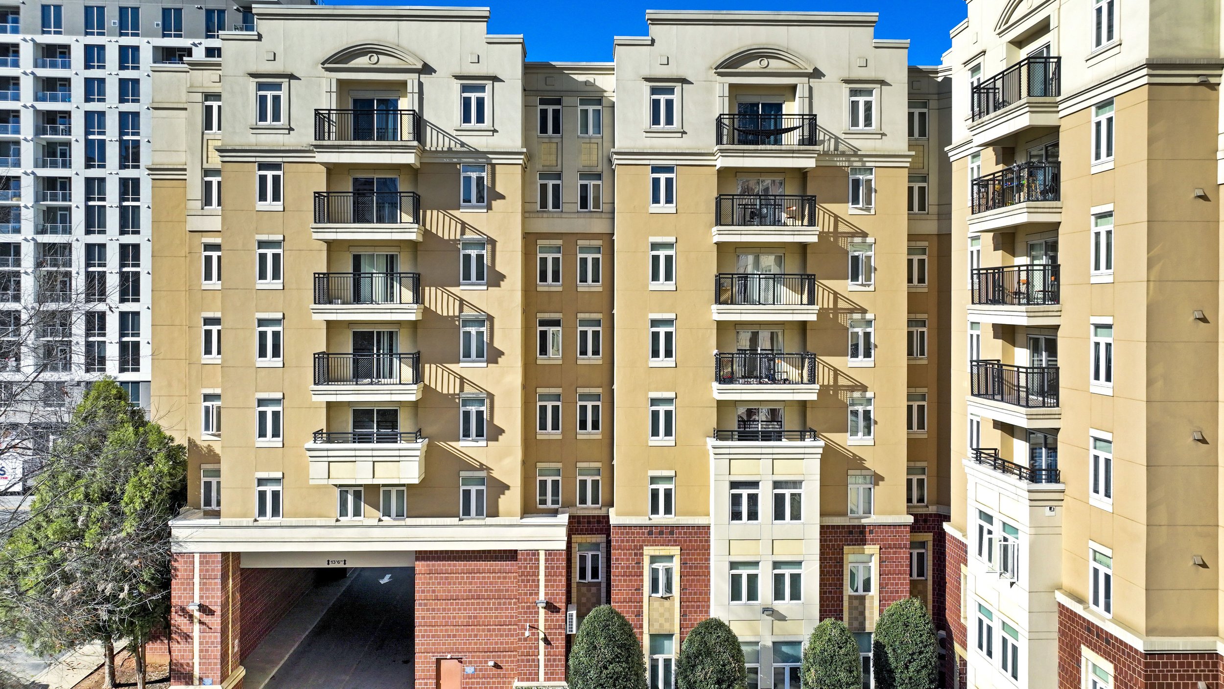 Front view of a multi-story apartment building with beige and red brick exterior, featuring balconies with black railings, white-framed windows, and an underground parking entrance at the ground level.