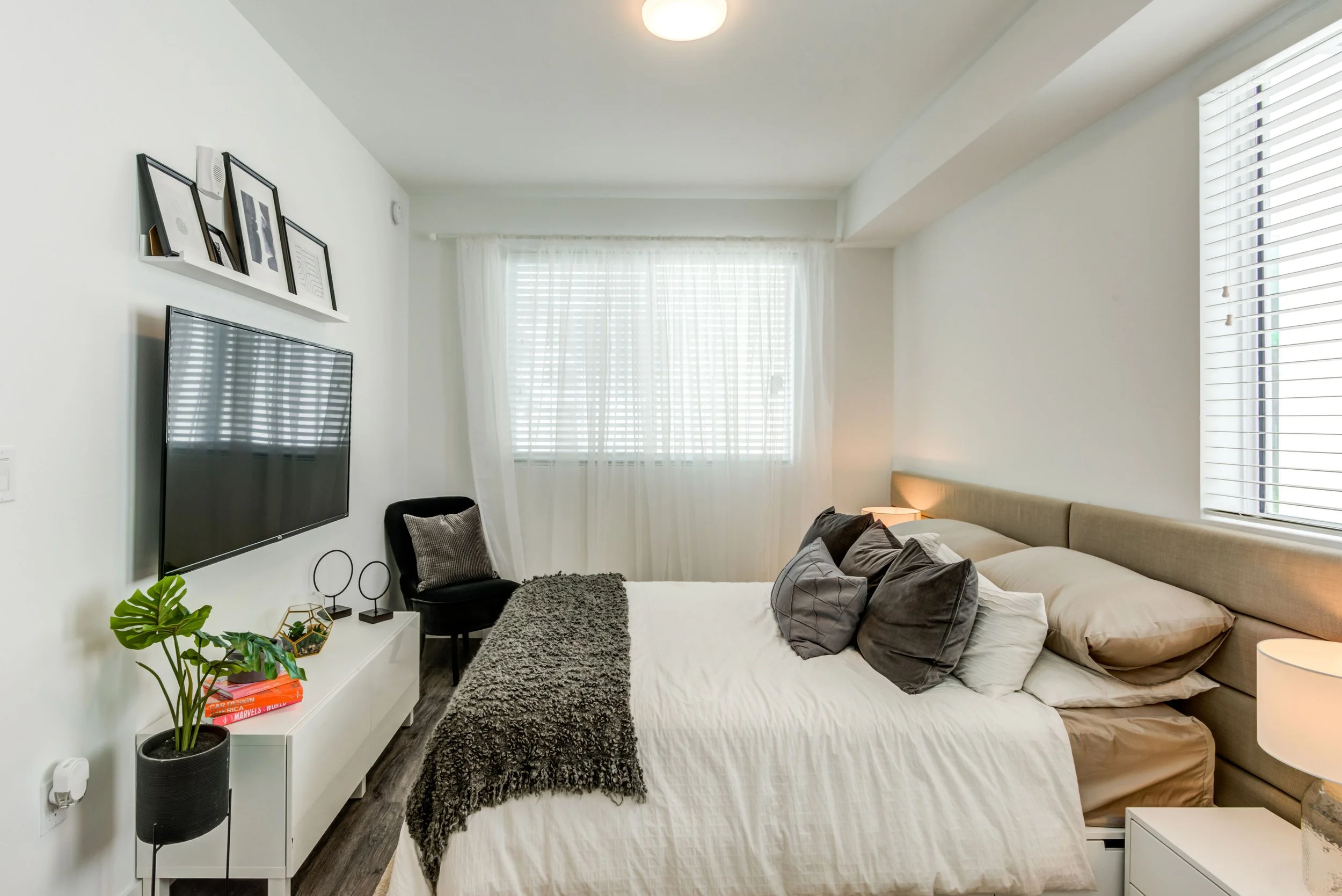 Modern bedroom with large window, beige bed, multiple pillows, white nightstands, black armchair, wall-mounted TV, white shelf with pictures, potted plant, and decorative items.