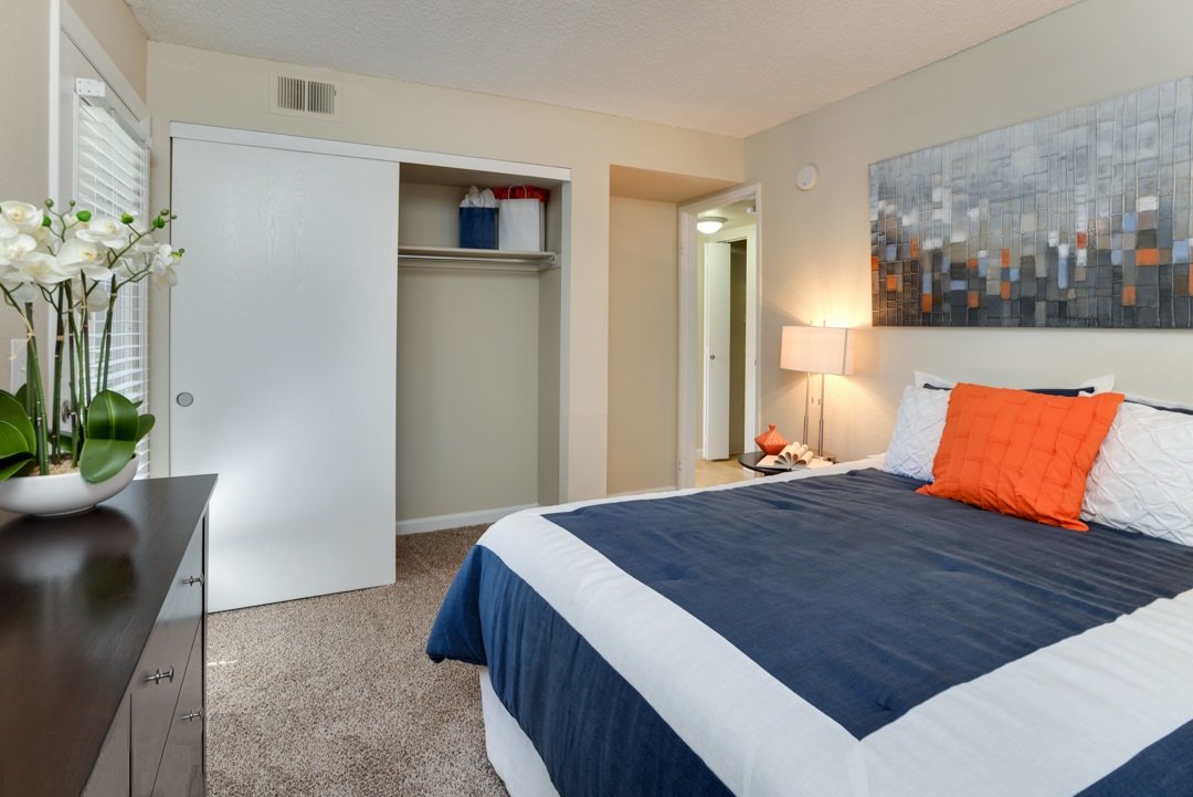 A bedroom with a bed, orange throw pillow, white and blue bedding, a nightstand with a lamp, artwork on the wall, a dresser topped with a plant, a closet with sliding door, and an open door leading to a hallway.