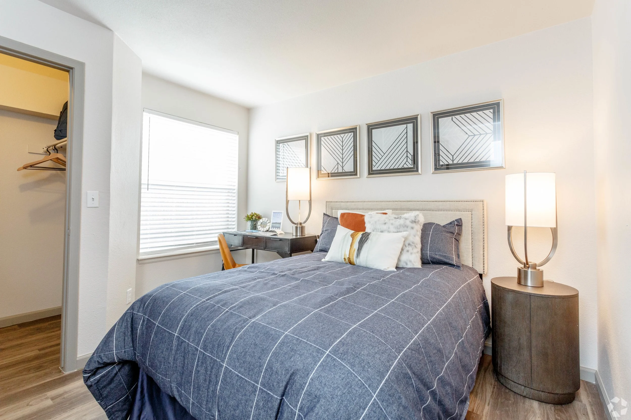 Bedroom with bed, nightstand, lamps, framed art, window with blinds, desk, orange chair, and closet with open door and hanging clothes.