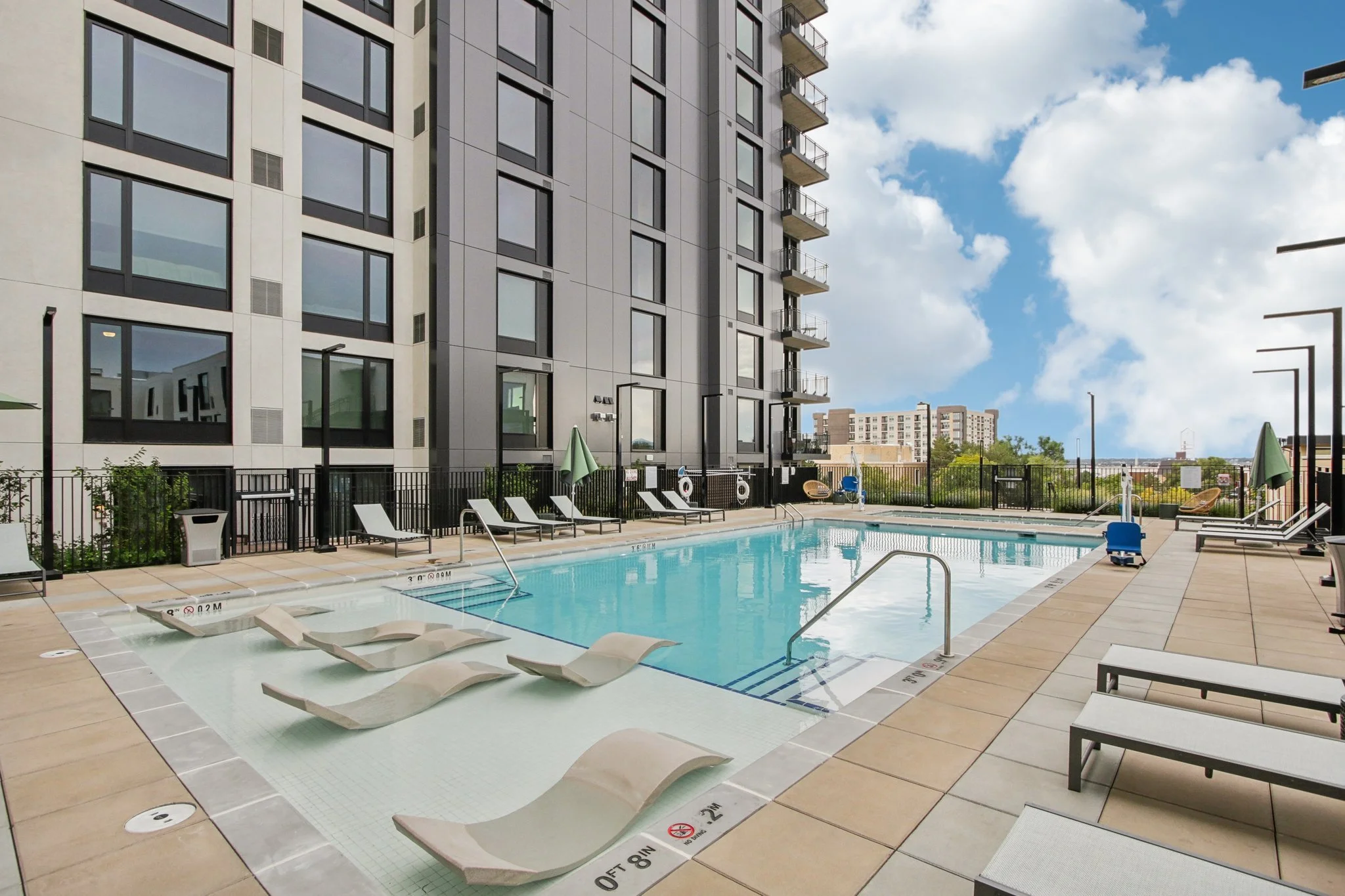 Outdoor rooftop swimming pool surrounded by lounge chairs, umbrellas, and a high-rise building structure with a cloudy sky in the background.