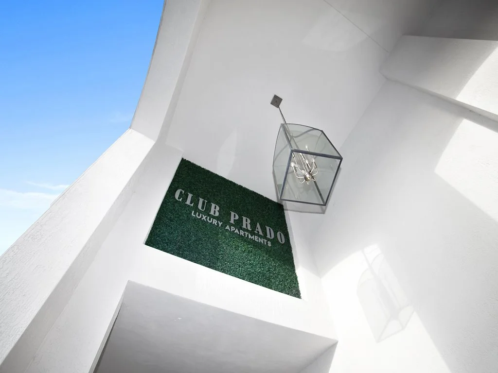 View of a white building corner with a green artificial grass square sign that reads 'CLUB PRADO LUXURY APARTMENTS', a modern glass lantern hanging from the ceiling, and a clear blue sky in the background.
