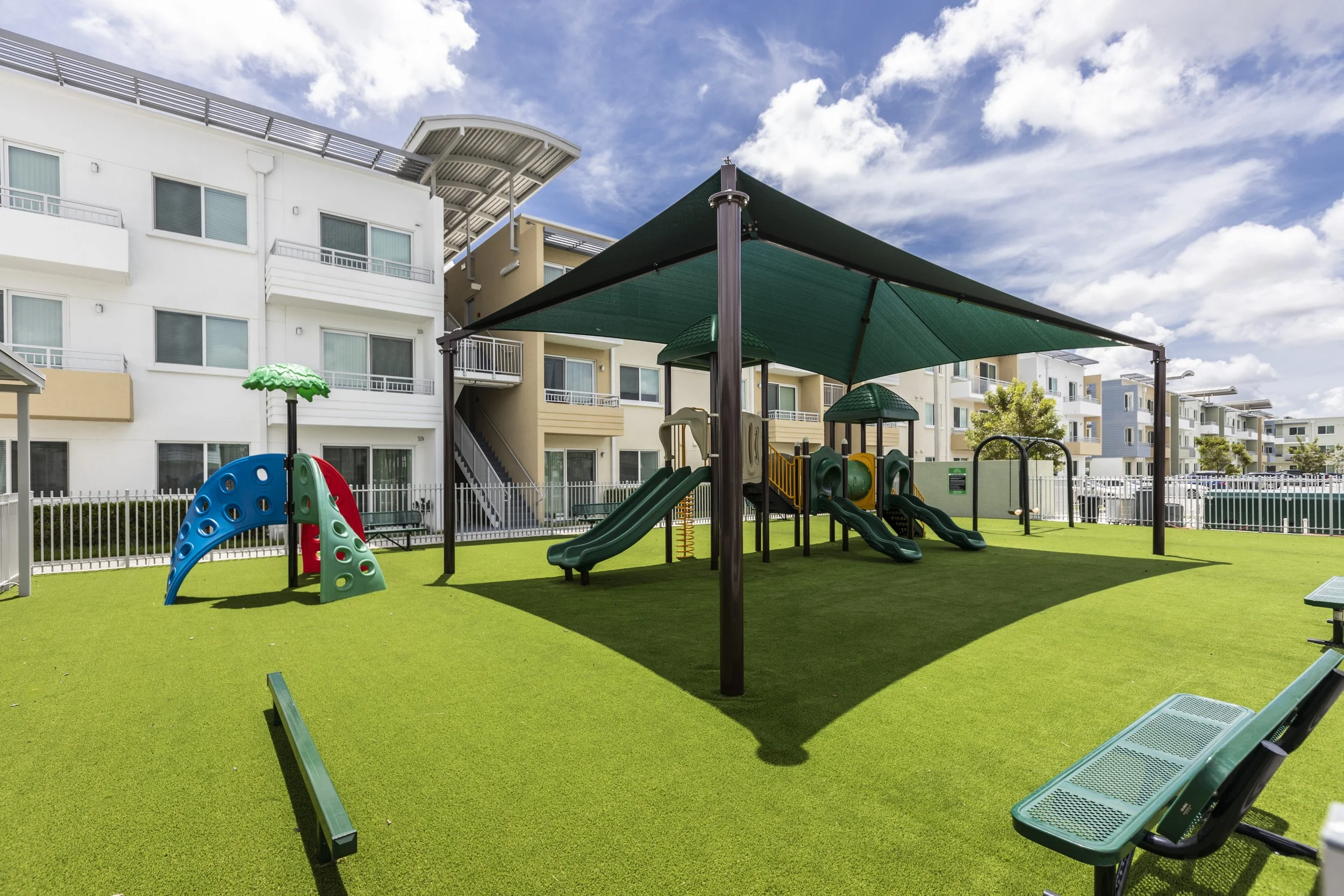 Public playground area with slides, climbing structures, shaded area, and benches surrounded by apartment buildings.