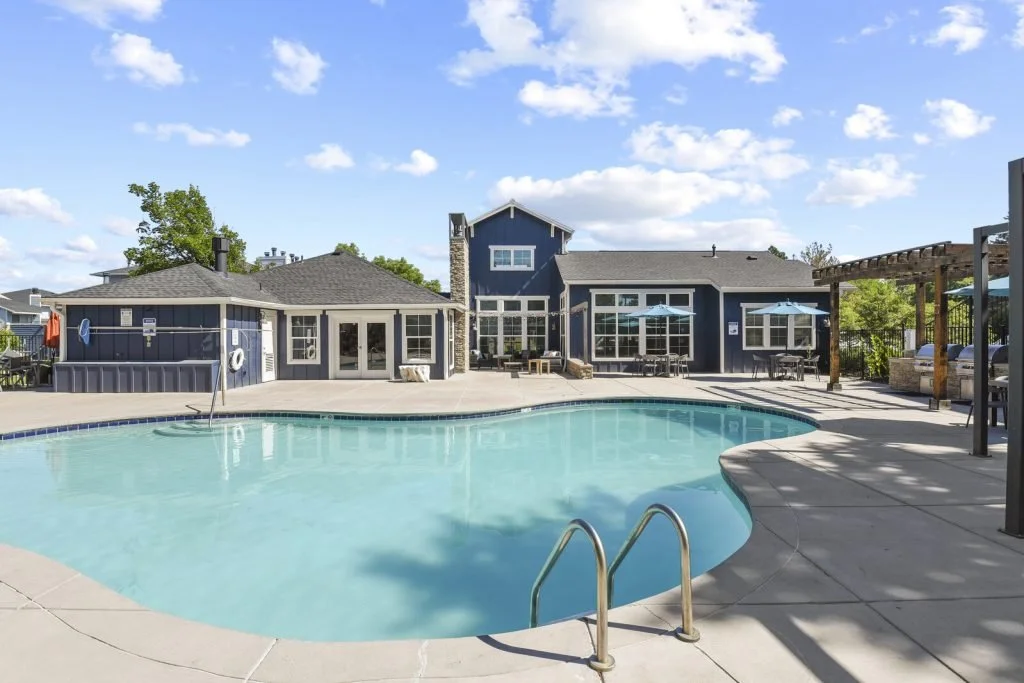 Community swimming pool with surrounding patio, lounge chairs, shaded areas with umbrellas, and a blue house with large windows in the background, under a partly cloudy sky.