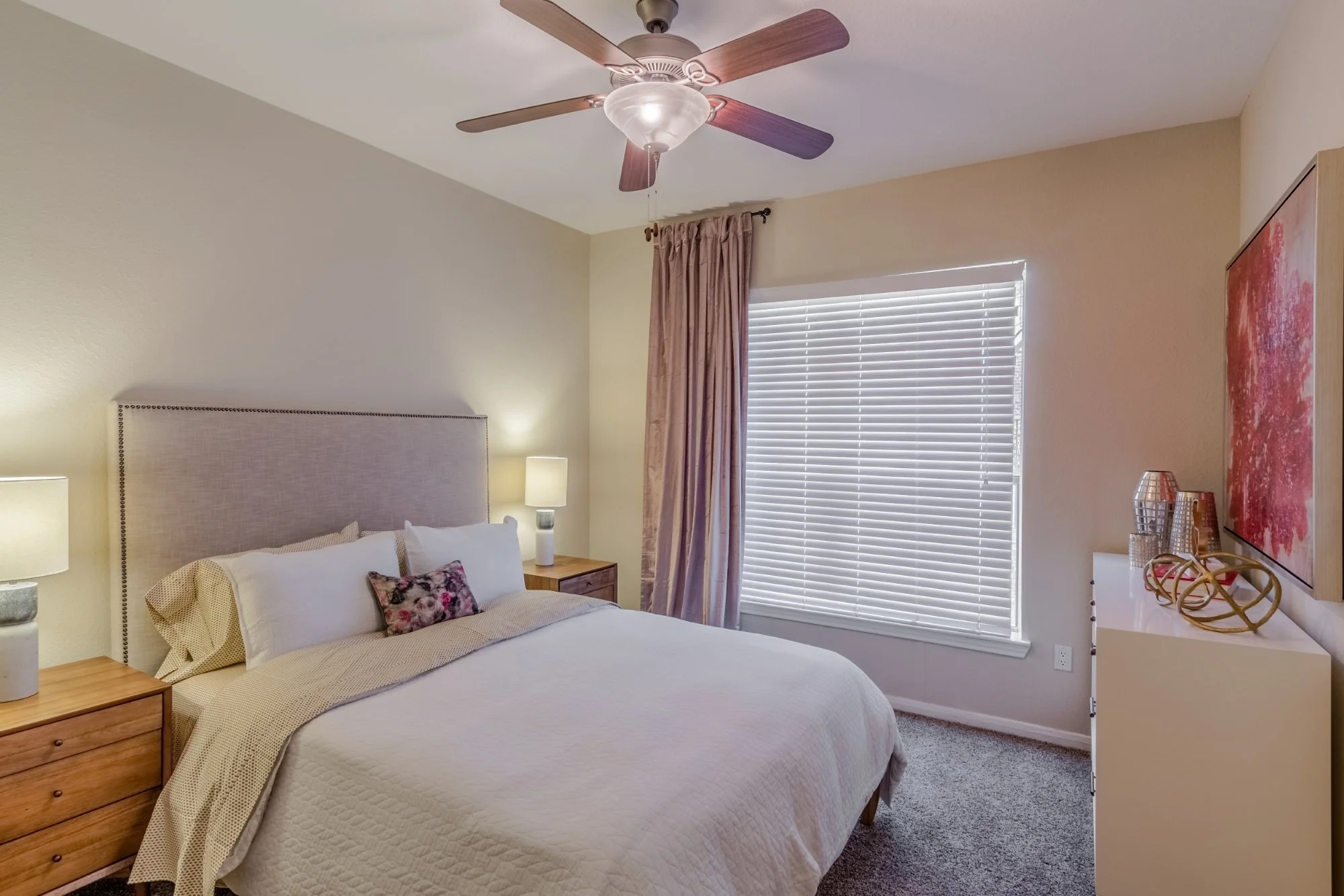 A bedroom with a bed, nightstands, lamps, window with blinds and curtains, ceiling fan, and wall art.