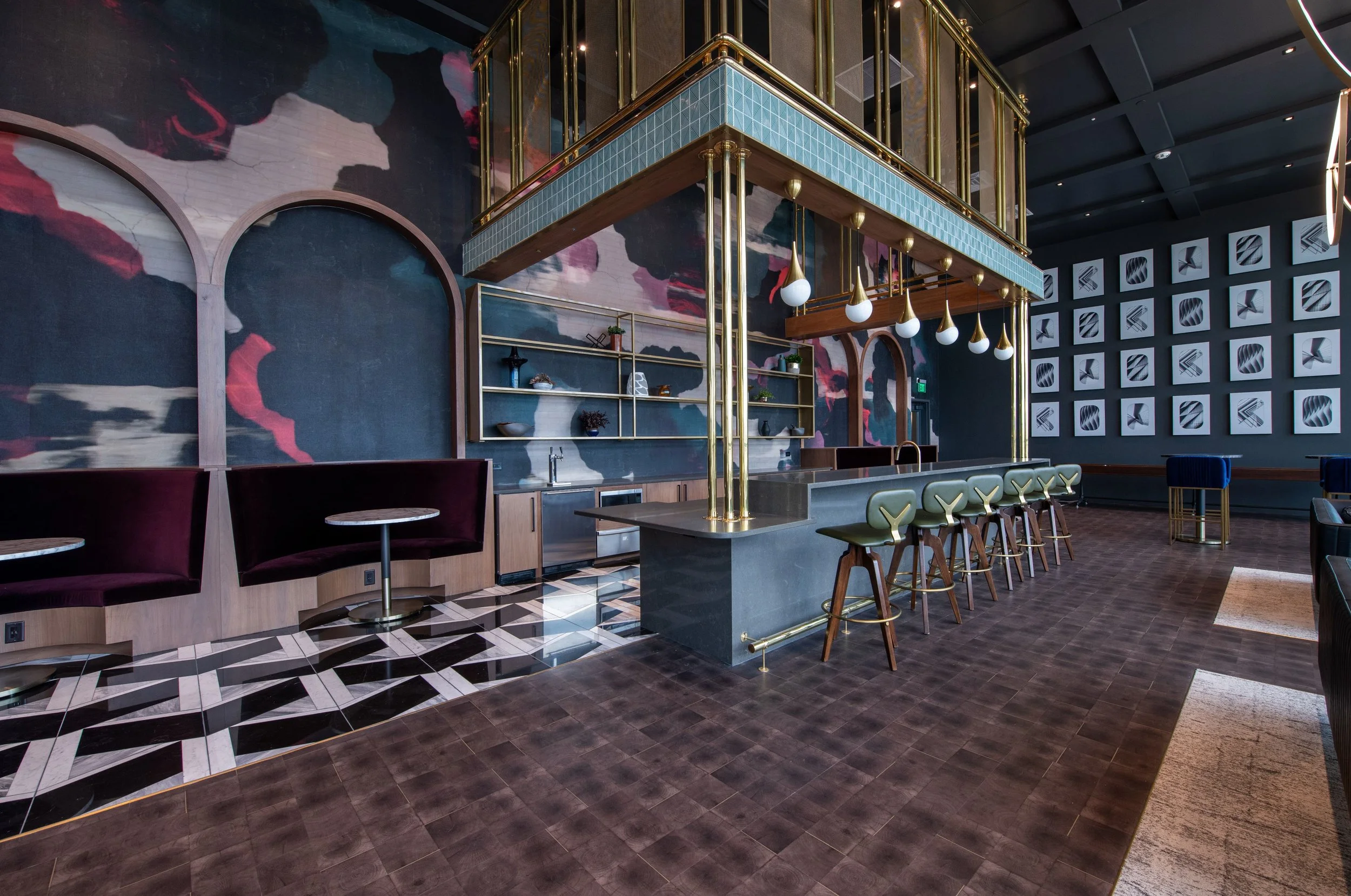 Modern, stylish bar interior with dark wood flooring, art wall, and gold accents, with a large bar counter surrounded by green and cream bar stools.