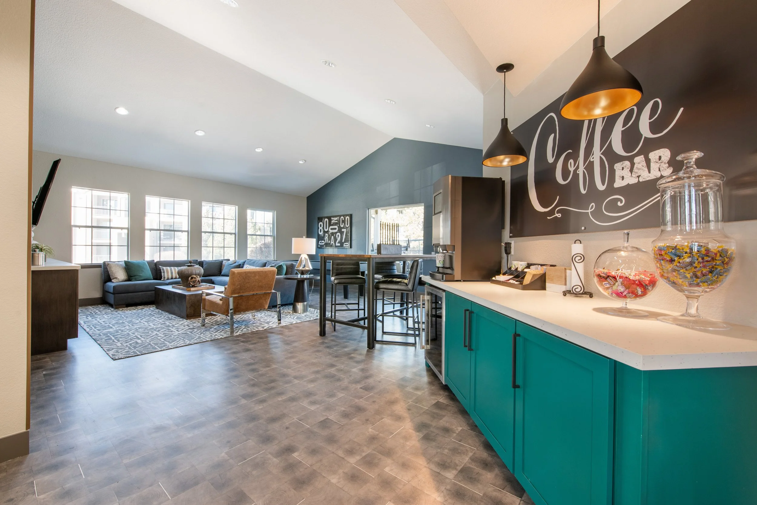 Indoor lounge area with large windows, teal cabinets, black bar stools, and a black chalkboard wall with white chalk writing that says 'Coffee Bar'.