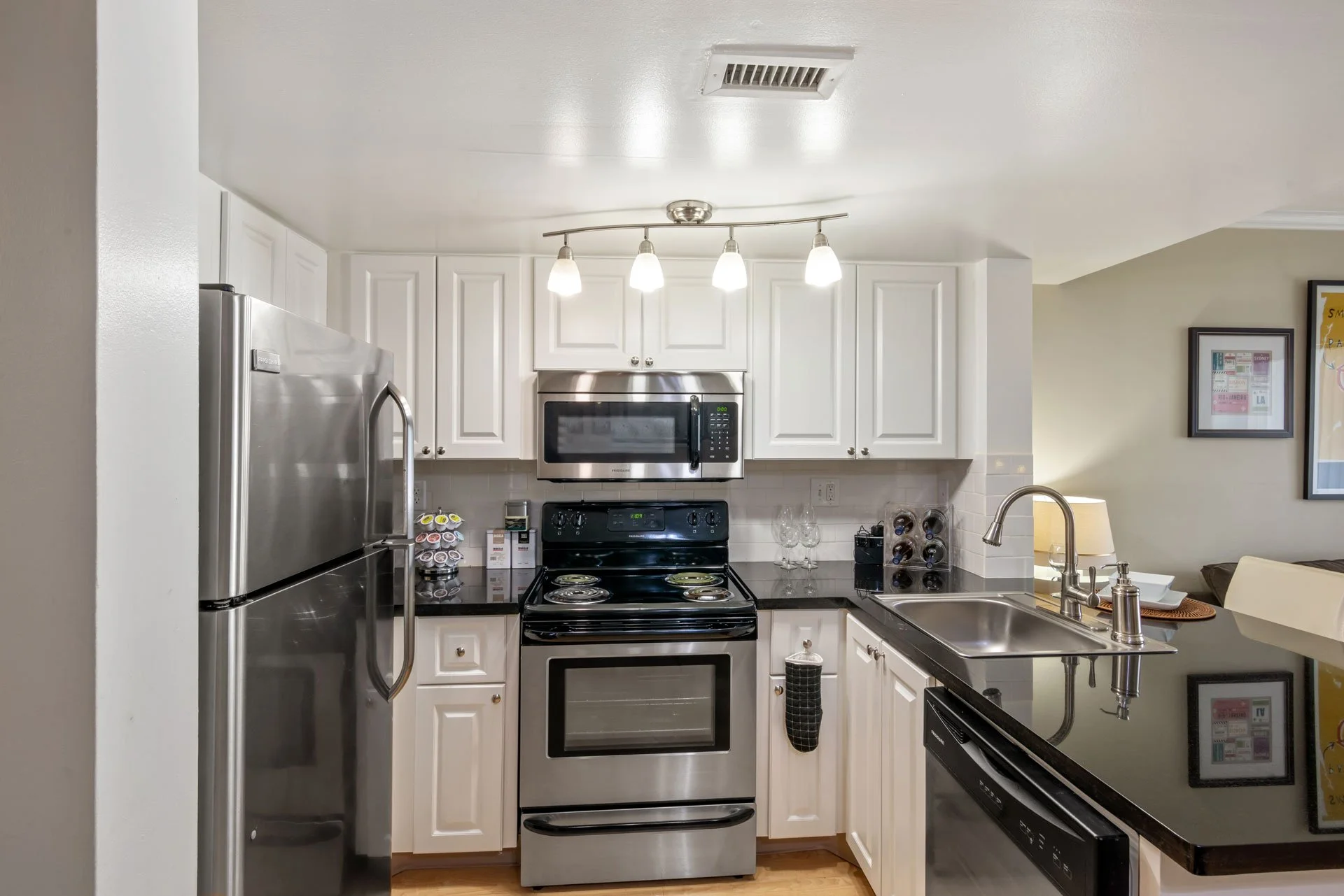 Kitchen with white cabinets, black countertops, stainless steel refrigerator, microwave, oven, and sink, with wine glasses and a wine rack on the counter.