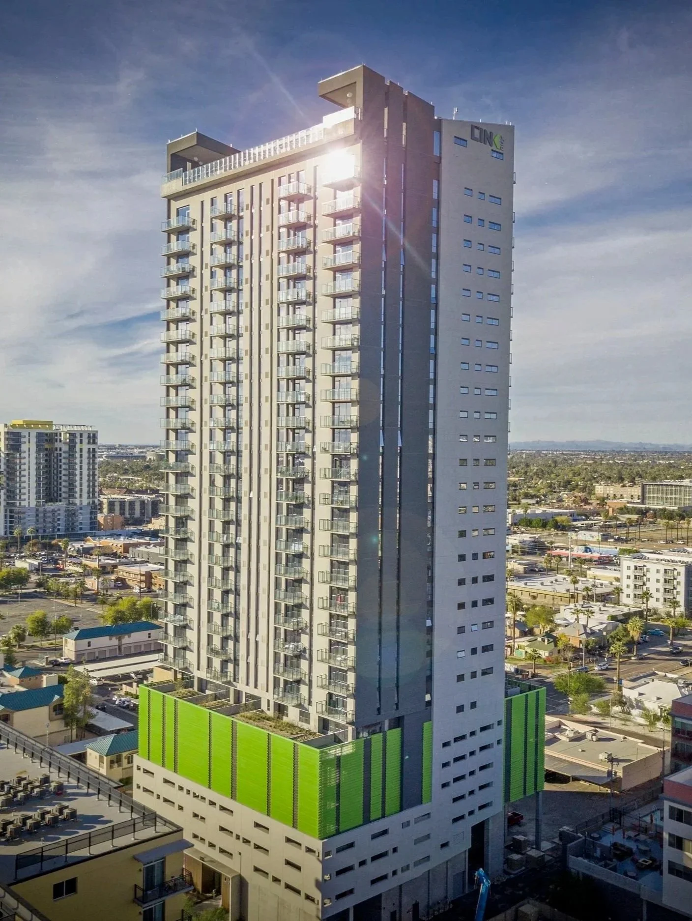 A tall modern high-rise residential building with numerous balconies and large windows, featuring a green and white lower section, with a cityscape and clear sky in the background.