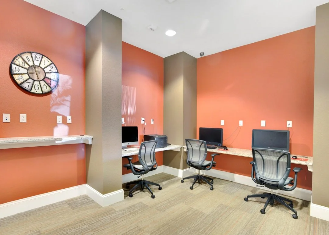 An office space with three desks, each with a computer and office chair, against rust-colored walls with beige accents and a large clock on the wall.