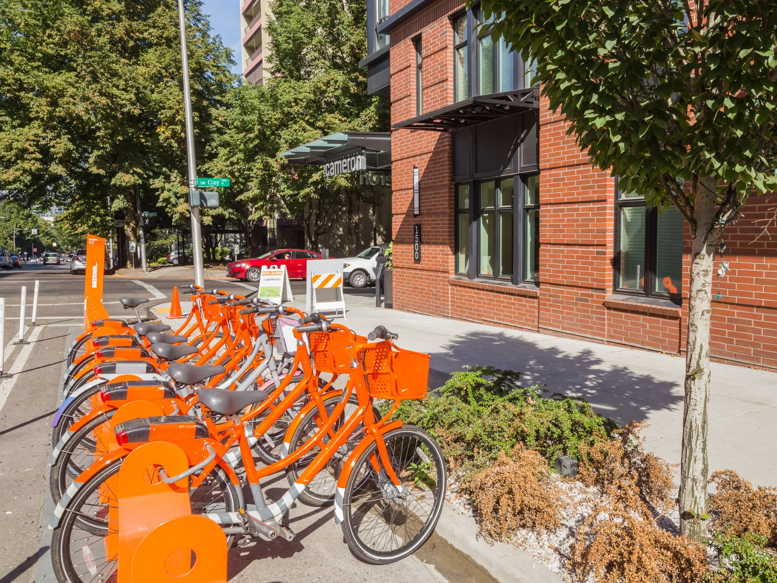 A line of orange and gray bicycles parked at a bike share station on a sidewalk, with a brick building, trees, and cars in the background on a sunny day.