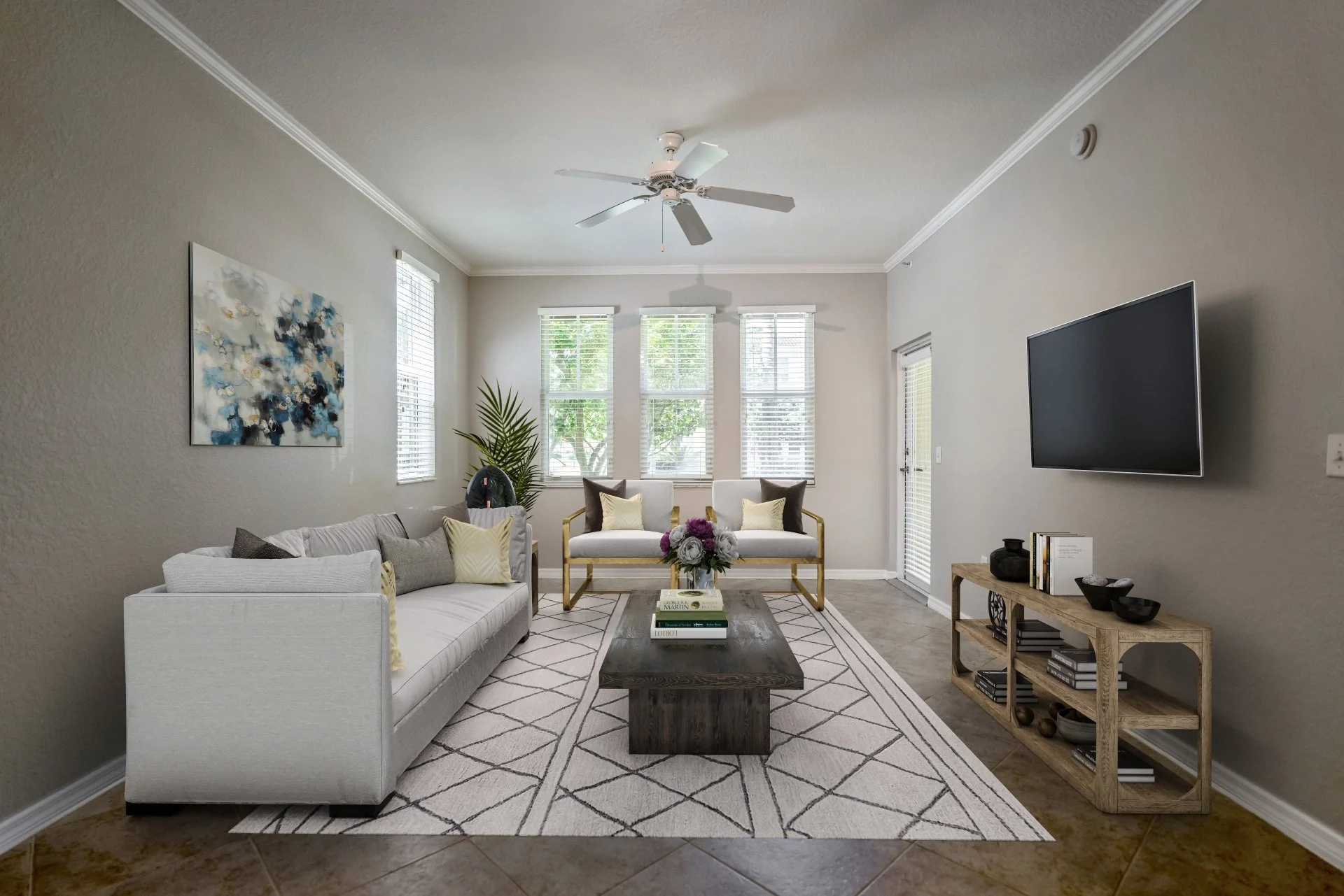 Living room with a white sofa, a wooden coffee table, and a wall-mounted TV. There are two armchairs with yellow and brown pillows, a potted plant, and a colorful bouquet on the coffee table. Windows with blinds are in the background, and the beige w