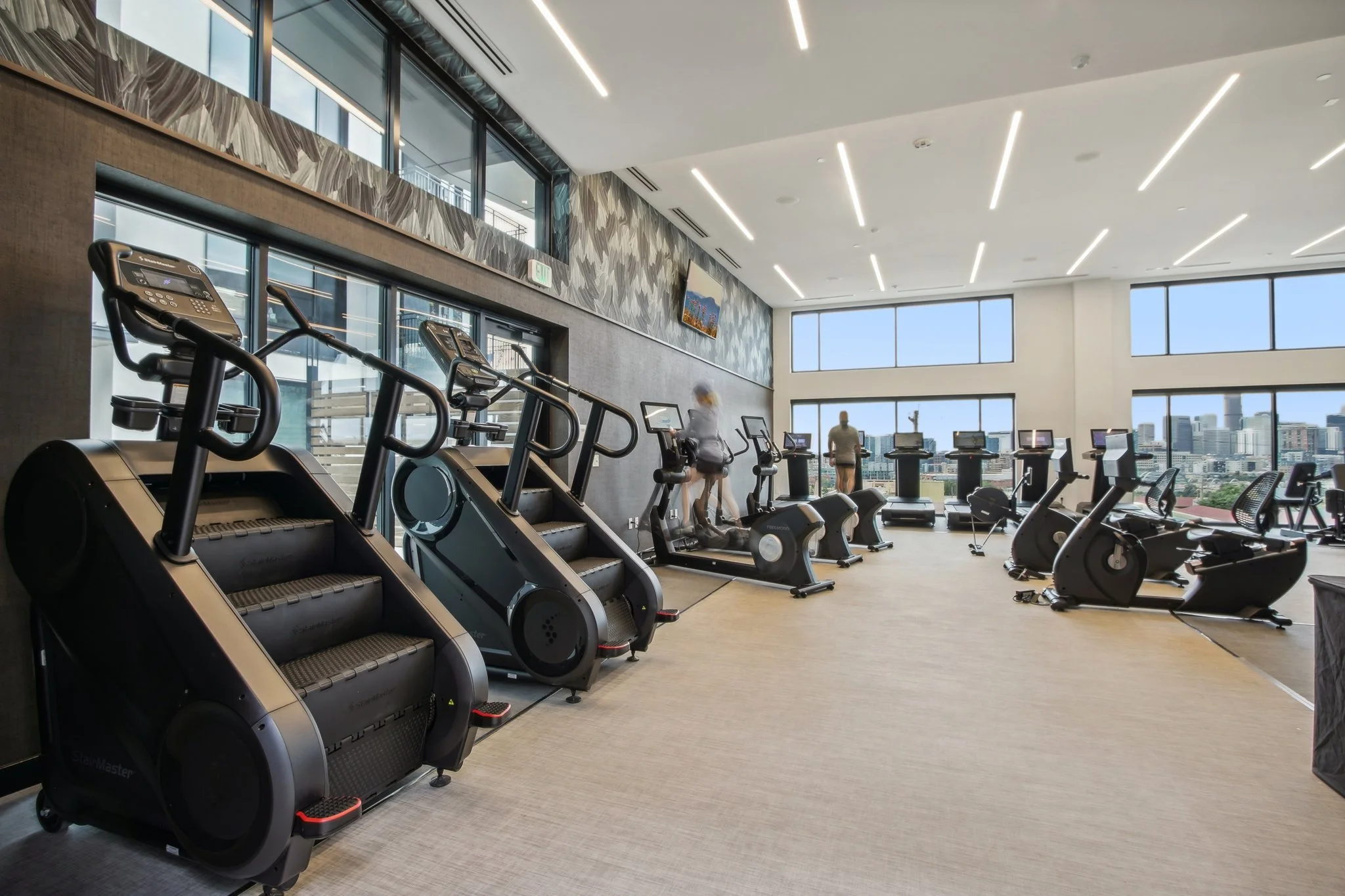 Modern gym with cardio equipment near large windows showing cityscape, people exercising.