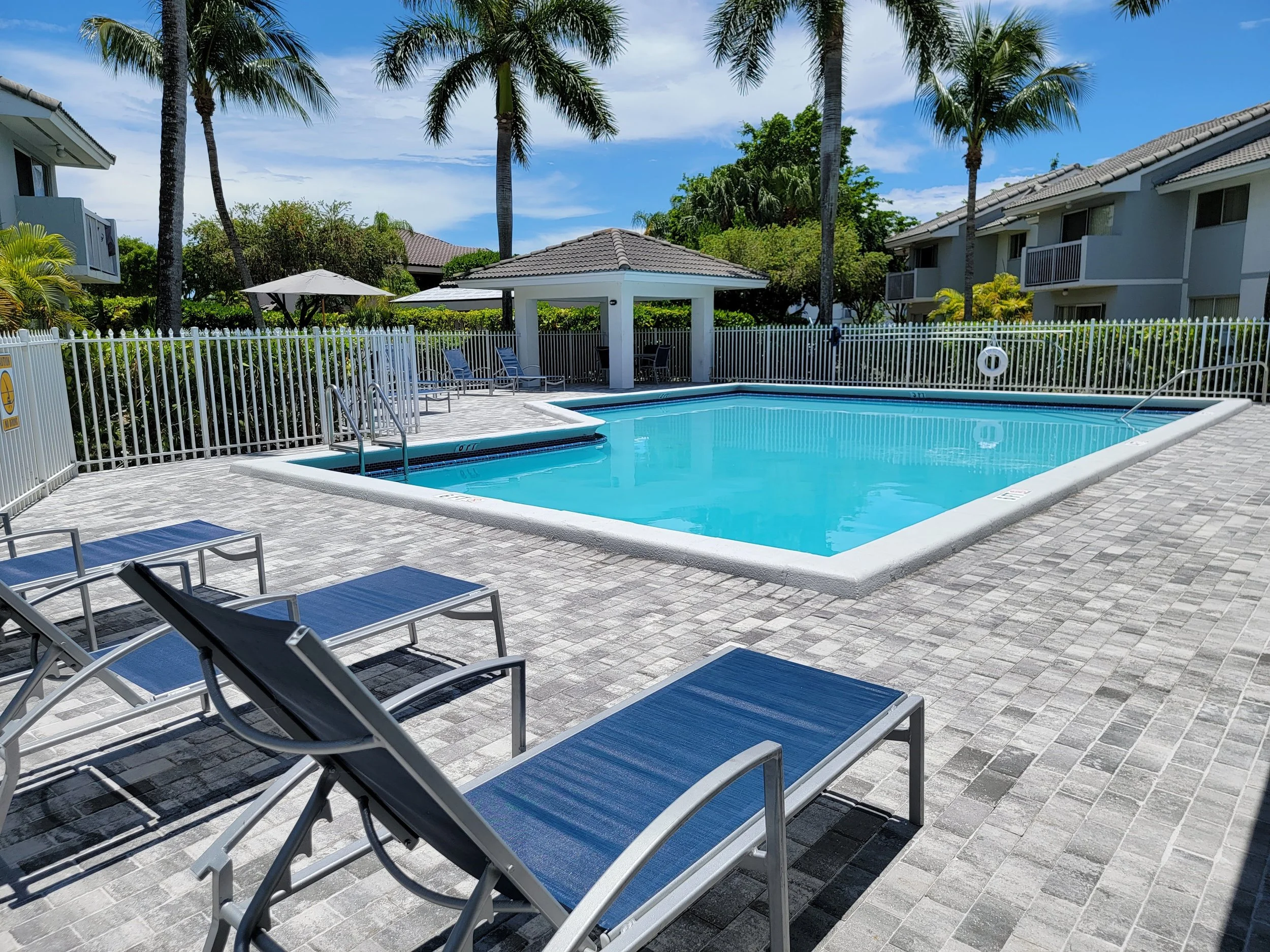 Empty outdoor swimming pool with blue water, surrounded by a white fence, lounge chairs, a gazebo, and palm trees under a blue sky with some clouds, in a residential complex.
