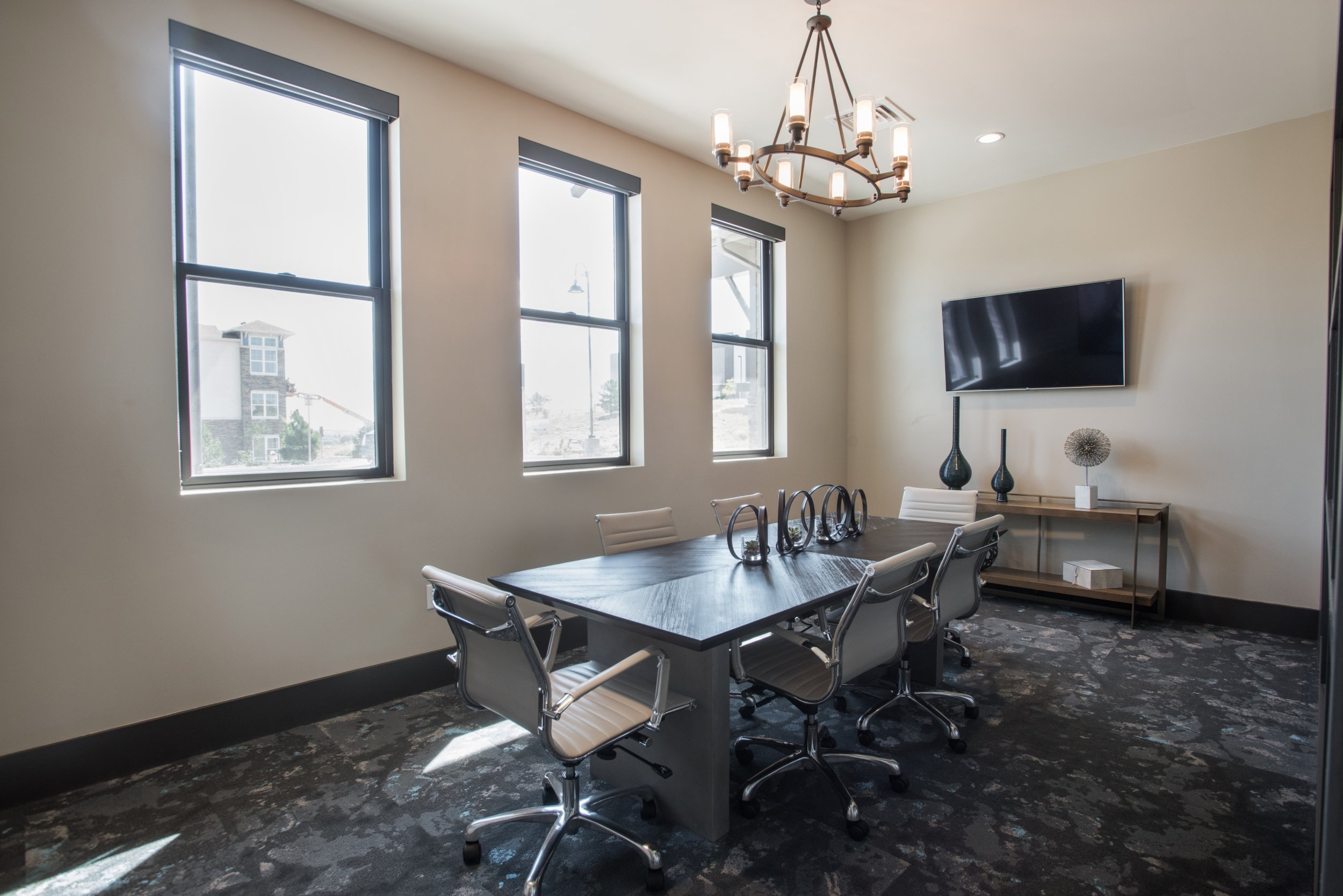 A modern conference room with a rectangular table surrounded by eight white office chairs. There are three windows on one wall, a chandelier hanging from the ceiling, and a flat-screen TV mounted on the wall with a side table below it decorated with 