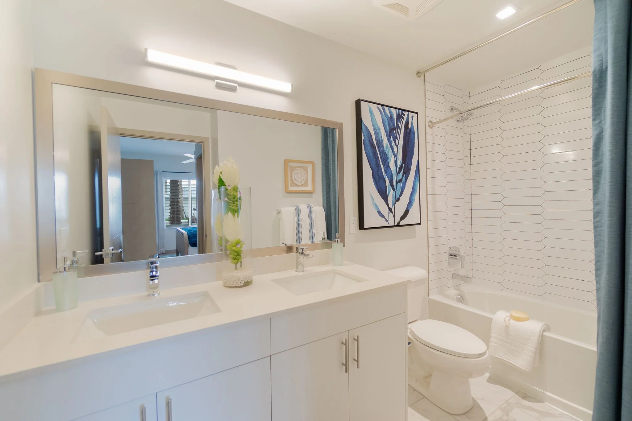Modern bathroom with white double sink vanity, large mirror, blue and green artwork, and a tub with a shower, towel, and soap.