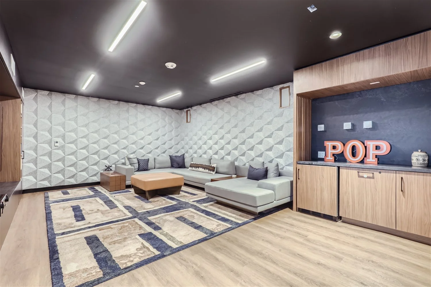 Modern living room with geometric patterned white wall, gray sectional sofa with pillows, geometric area rug, wooden side table, and a black wall with orange "POP" letters, wooden cabinetry, and decorative jar.