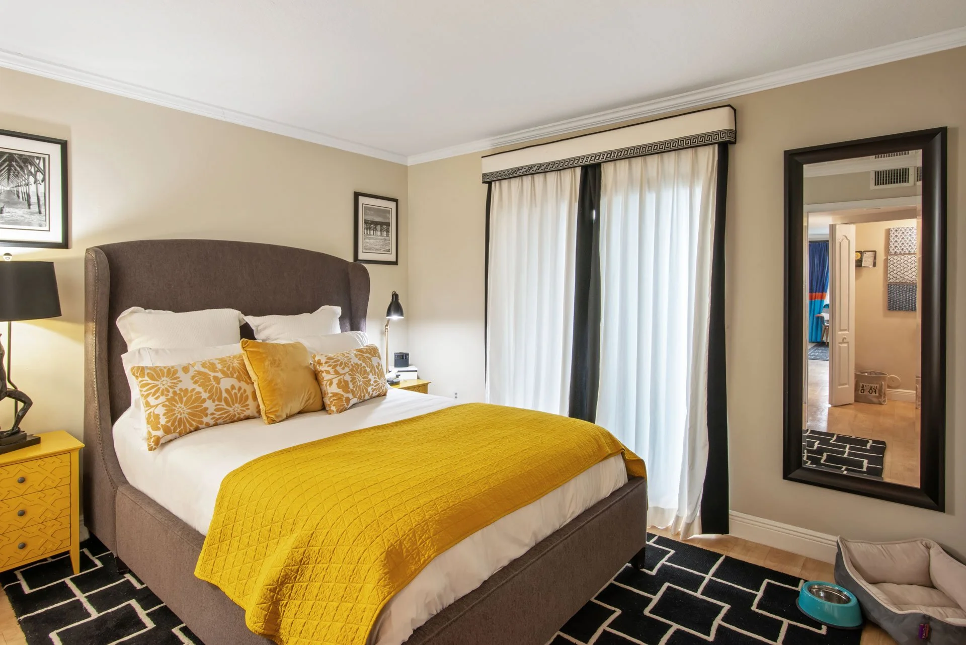 A bedroom with a large bed featuring a mustard yellow quilt and decorative pillows. There are black and white framed pictures on the cream walls, black and white curtains, and a full-length mirror on the wall. A nightstand with a lamp is next to the 