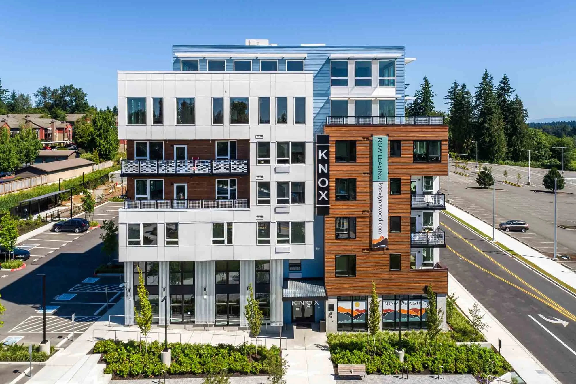 Modern multi-story apartment building with gray, white, and brown siding, featuring balconies with black railings, parking lot, and landscaped surroundings.