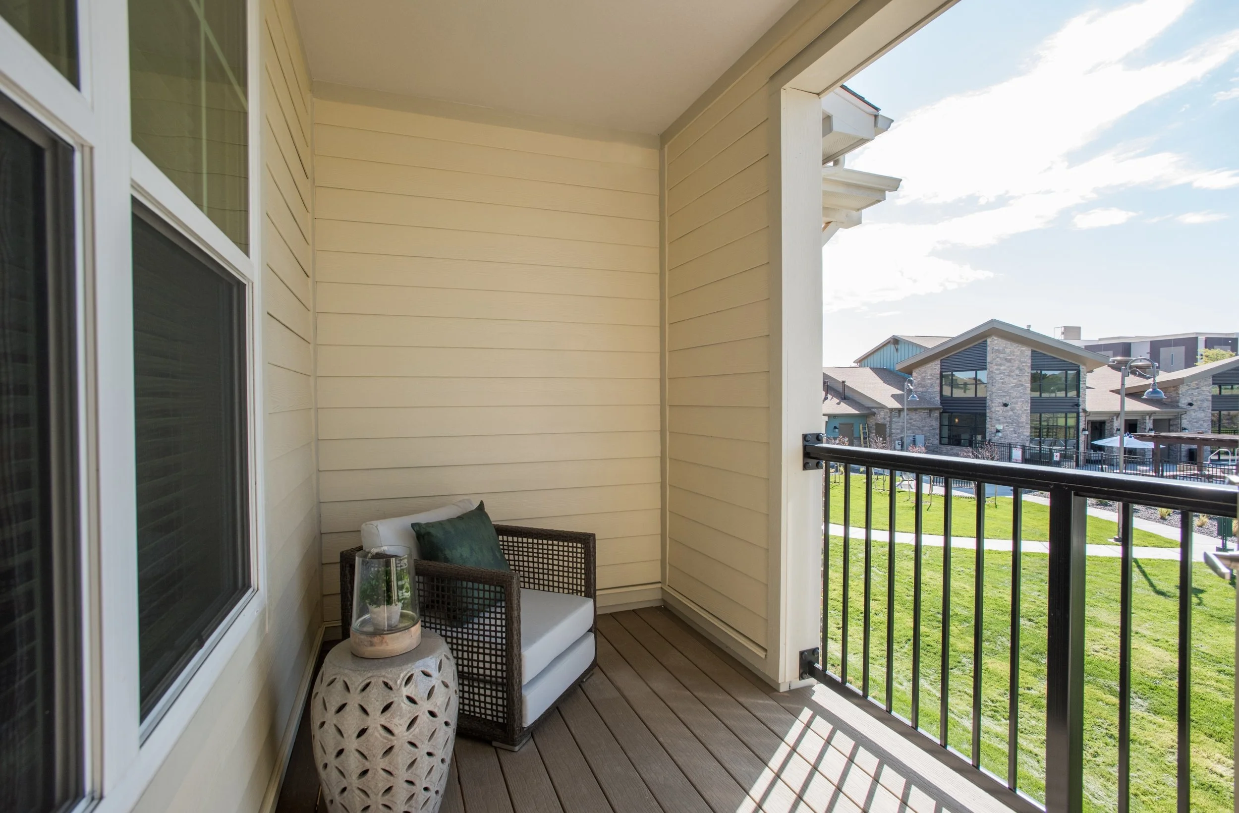 Small balcony with a wicker chair, a green pillow, a ceramic side table with a plant, overlooking a grassy area and modern neighboring buildings.