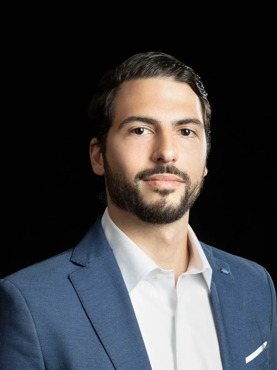 Portrait of a man in a dark suit, white shirt, smiling, with dark hair and a beard, against a dark background.