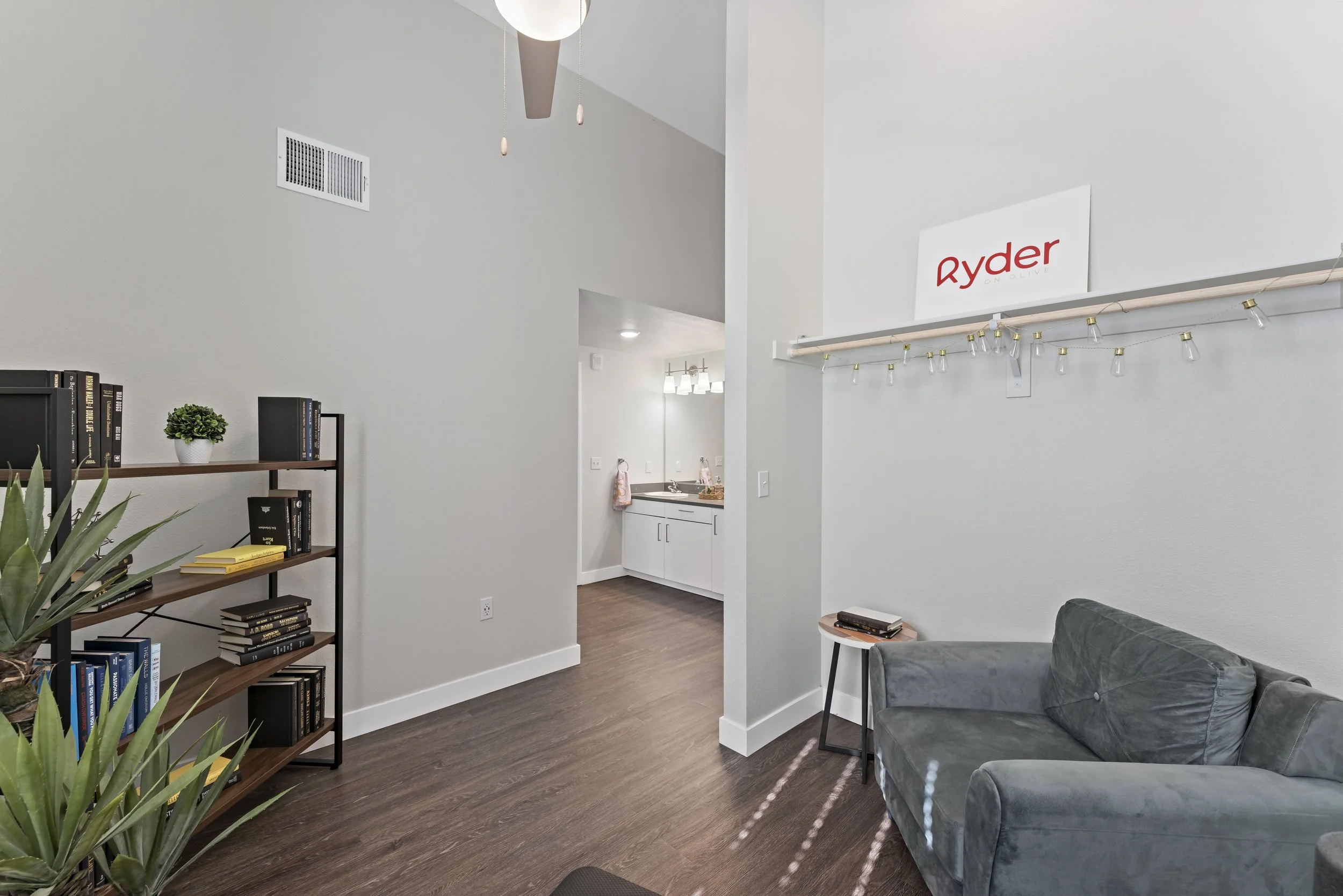 An interior living room with a dark gray velvet sofa, a small side table, a wooden bookshelf filled with books, a potted plant, and a sign on the wall that says 'Ryder'. The space has white walls, brown wood flooring, and is connected to a small kitc