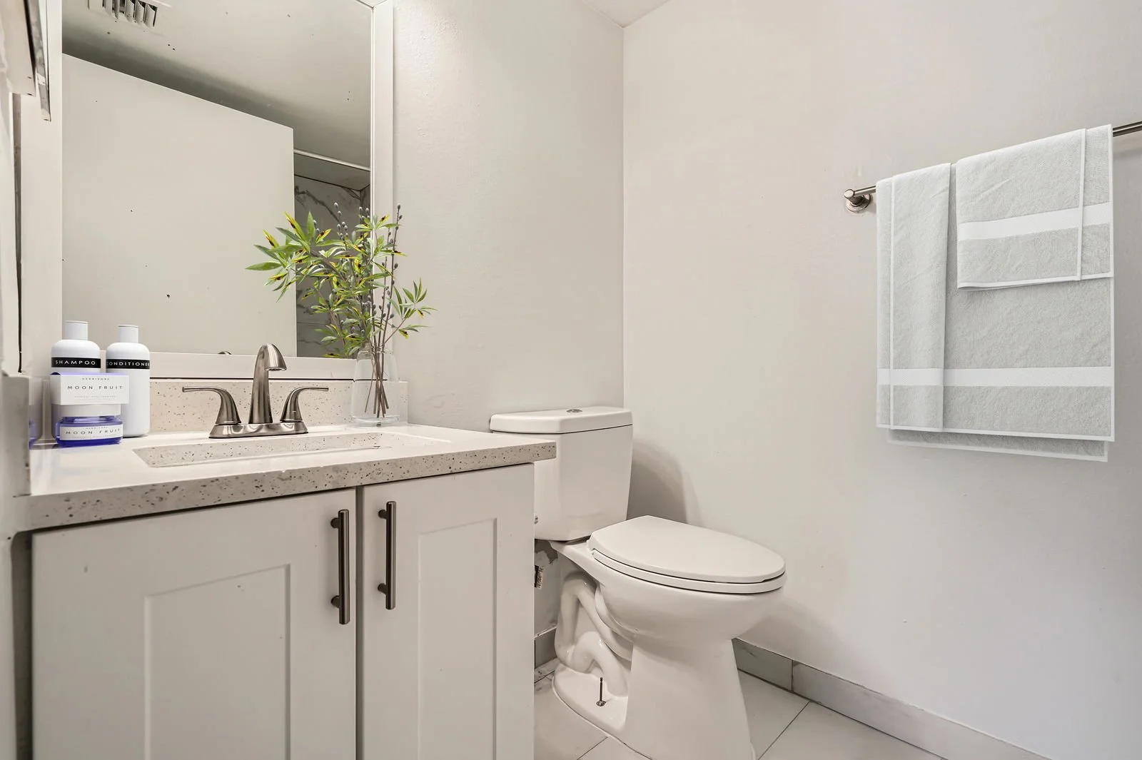 A modern bathroom with a white vanity, speckled countertop, and faucet. A potted plant and toilet are visible, with a mirror above the sink and a towel hanging on the wall.