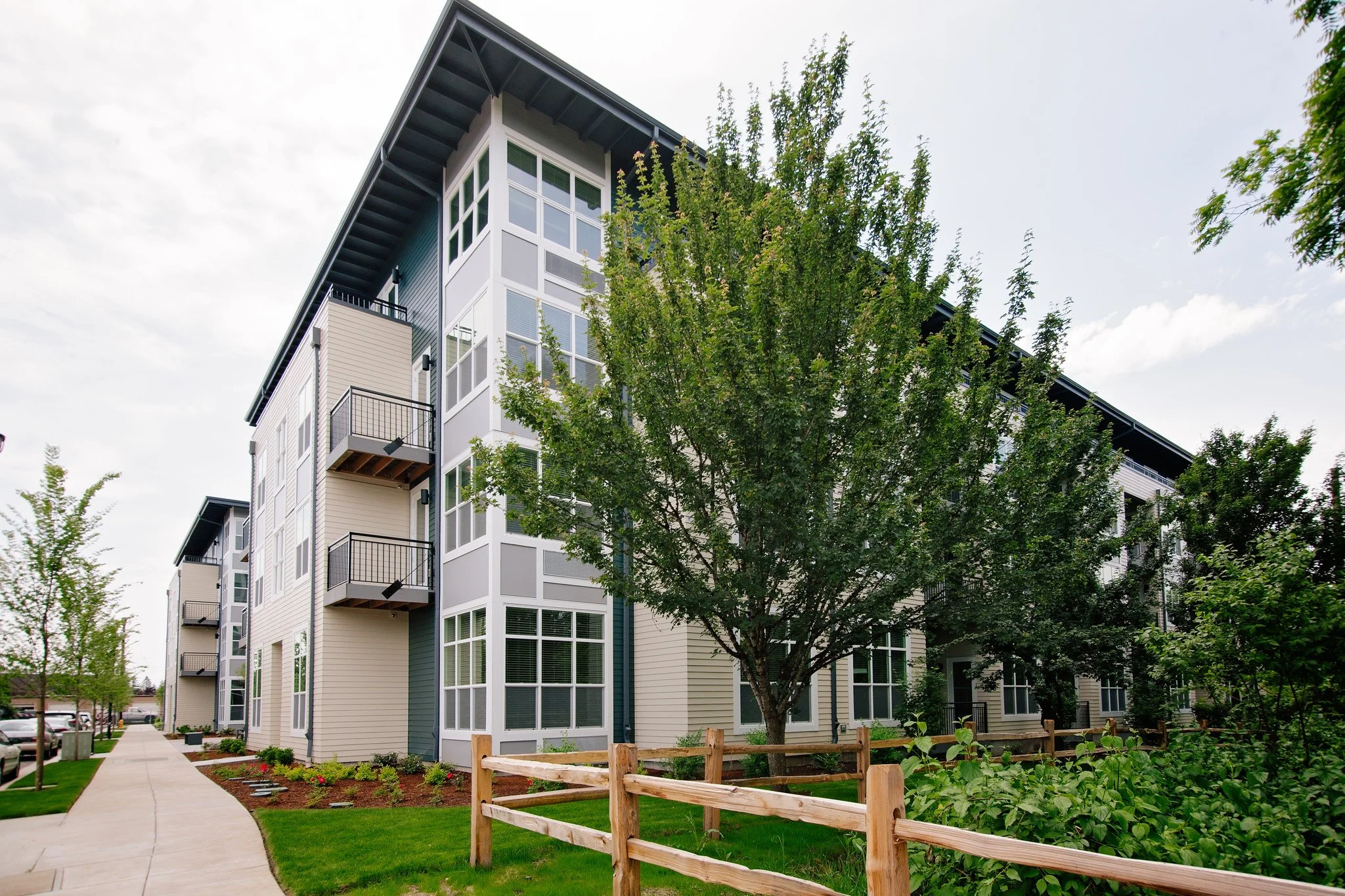 Modern apartment building with multiple balconies and large windows, surrounded by green trees, a wooden fence, and a sidewalk.