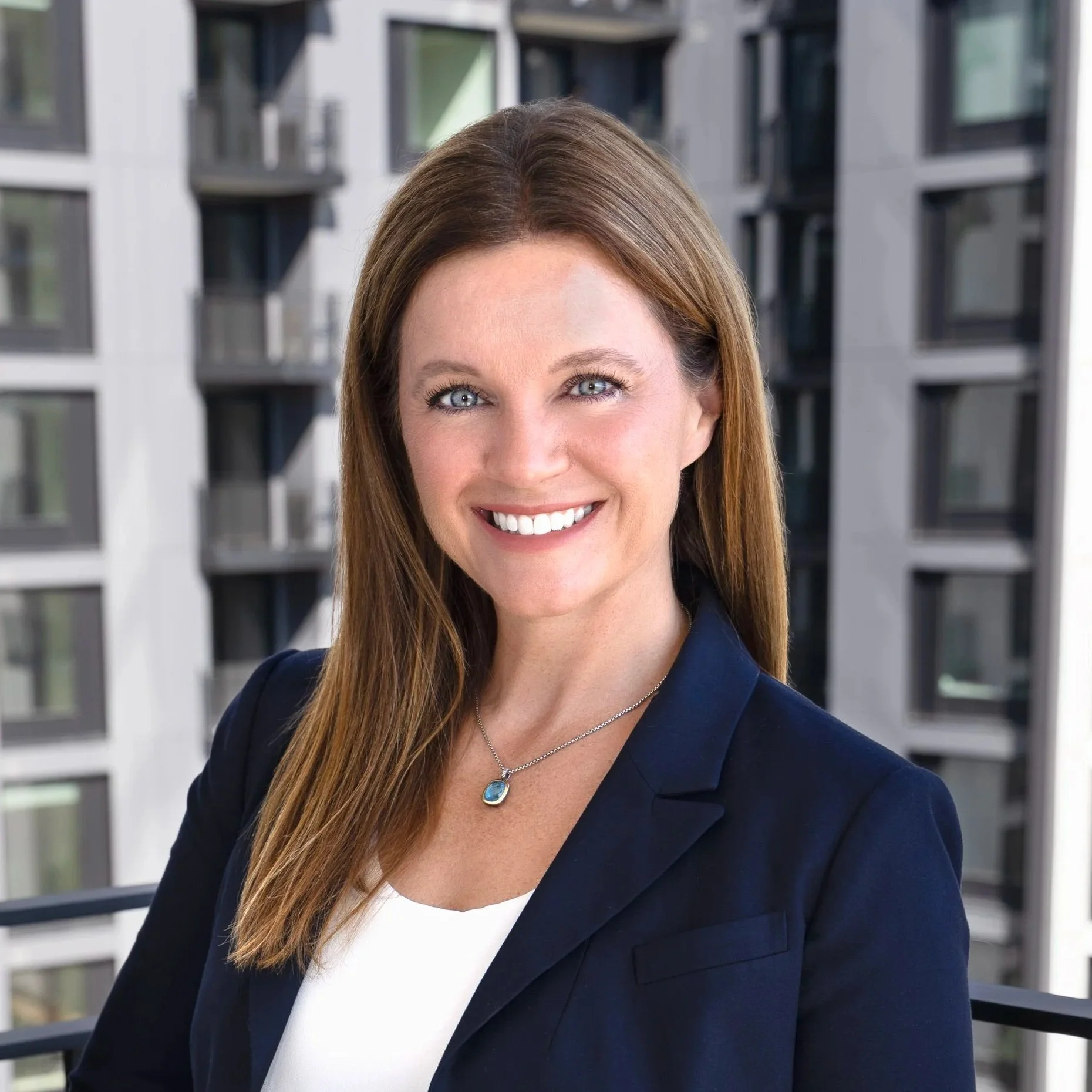 A woman with long red hair and blue eyes smiling, wearing a navy blazer and a silver necklace with a blue pendant, standing outdoors with a city apartment building in the background.