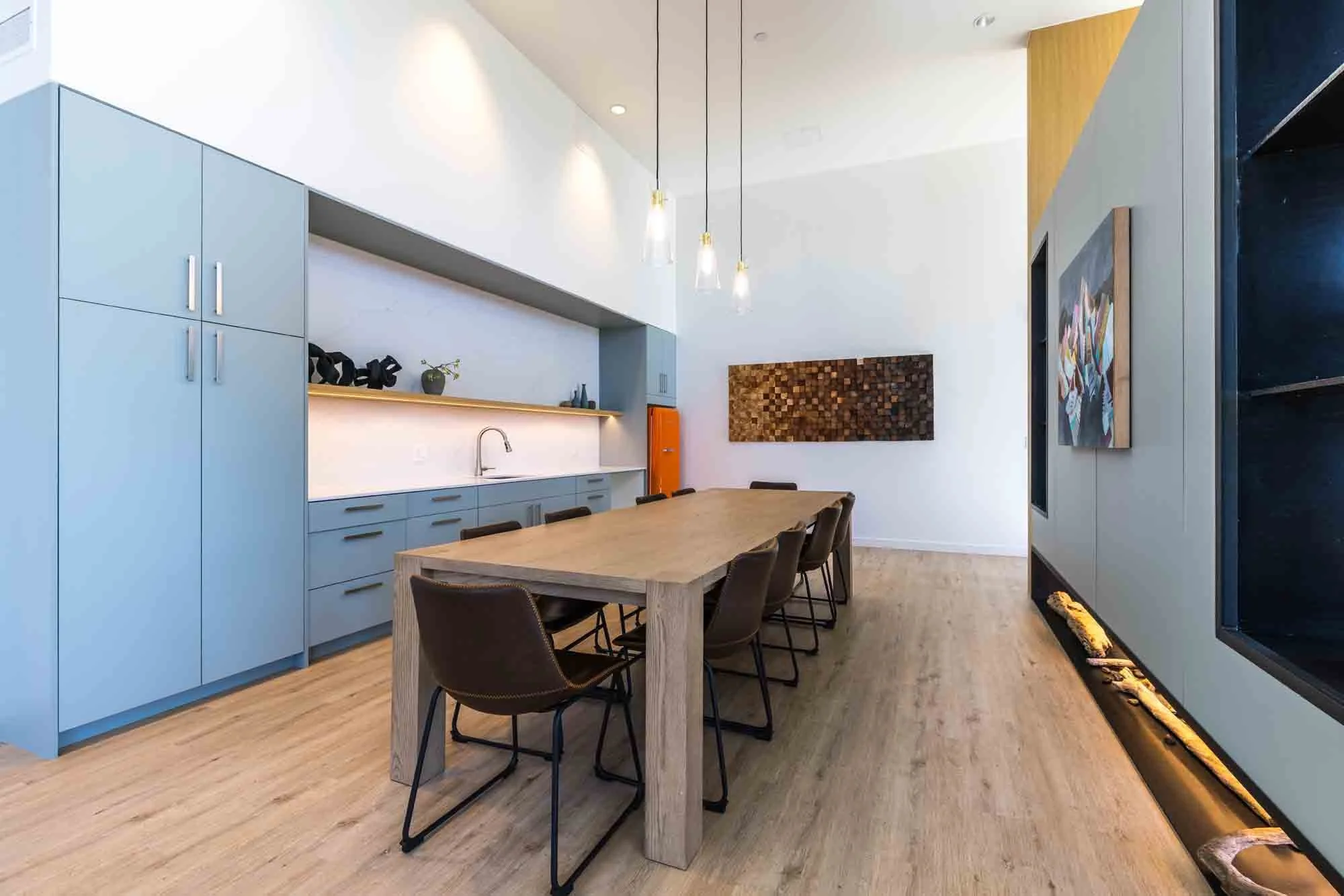 Modern kitchen with light blue cabinets, wooden dining table, six dark brown chairs, wood flooring, wall art, and pendant lighting.