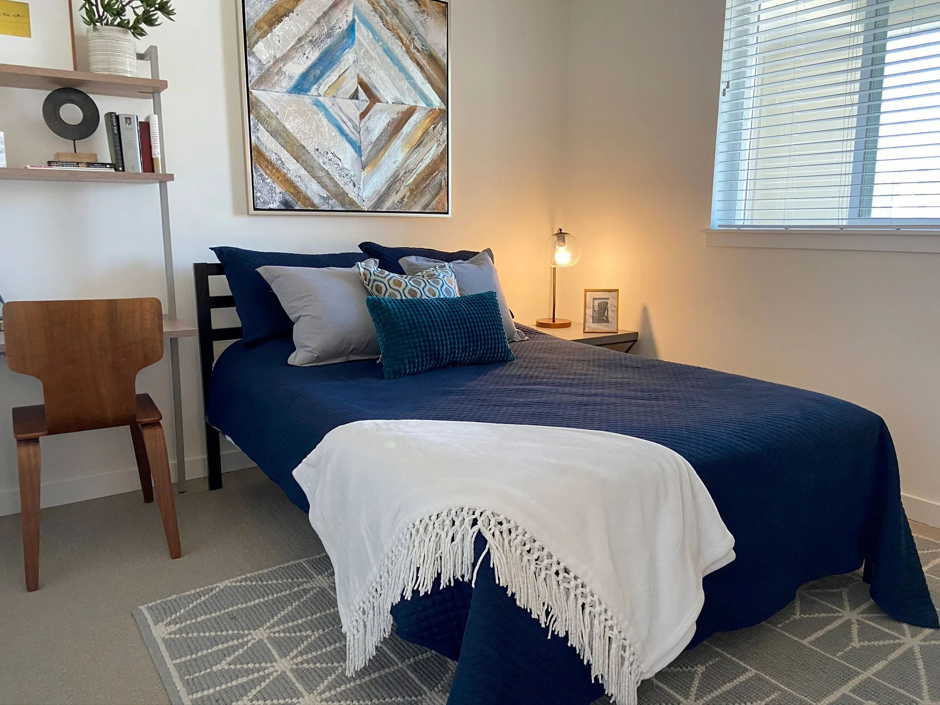 A neatly made bed with navy blue quilt and pillows, white throw blanket, bedside lamp, framed art, and a window with closed blinds in a modern bedroom.