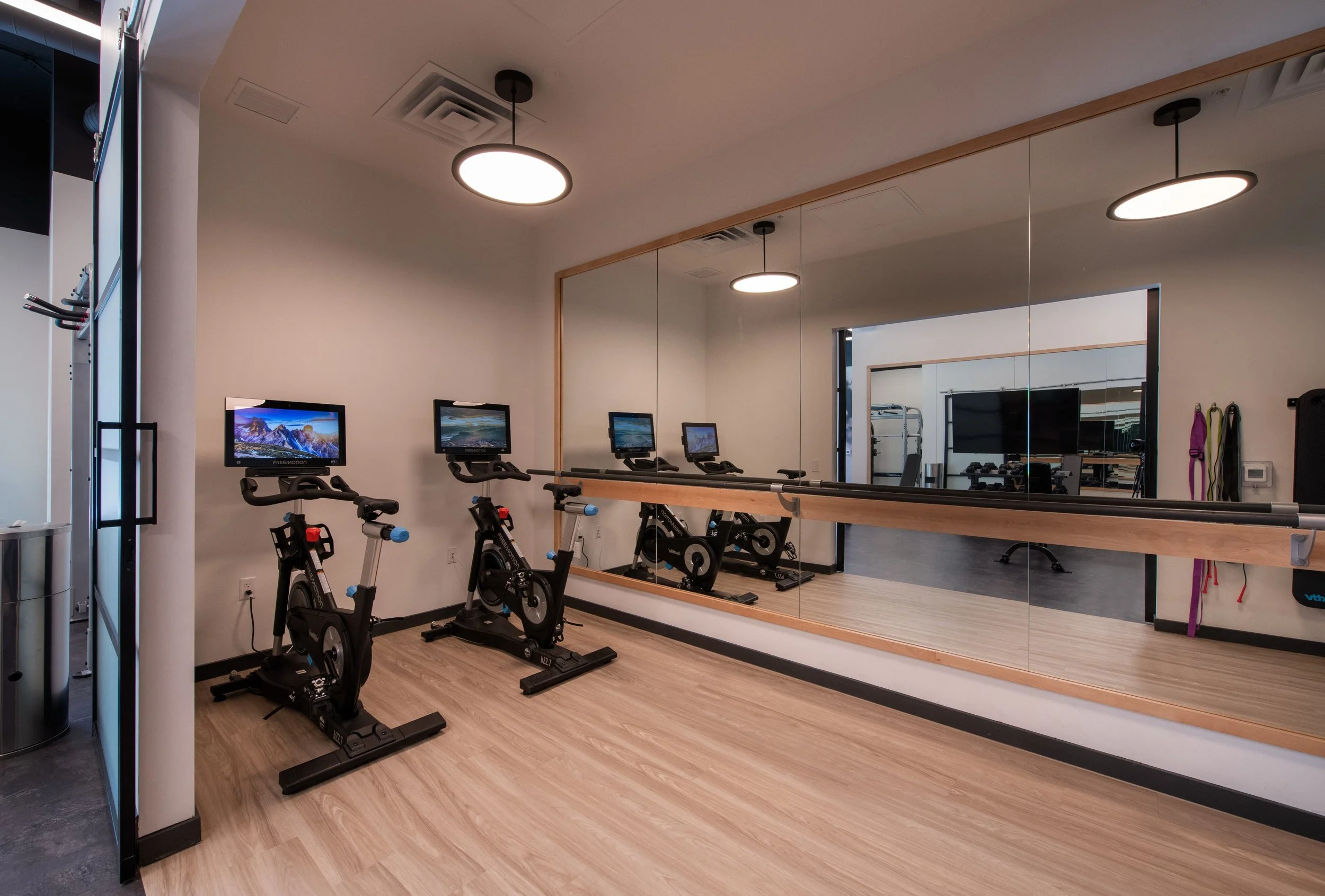 Indoor fitness room with three stationary exercise bikes, large mirror wall, and exercise equipment in the background.