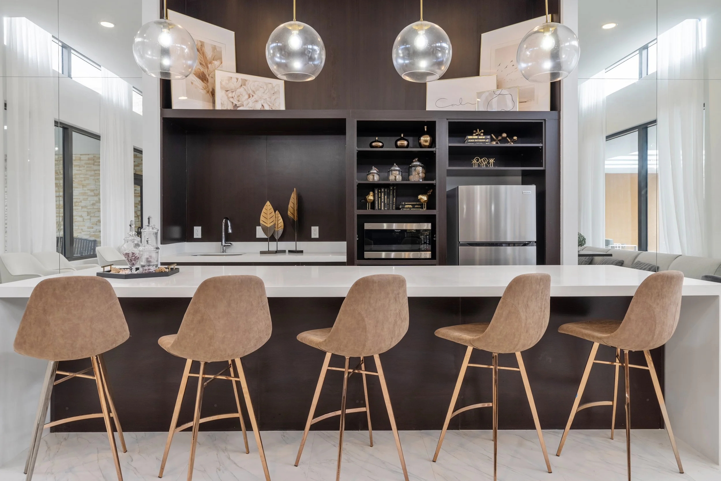 Modern kitchen with a white island counter, six tan barstools, dark wood cabinetry, and pendant lights.