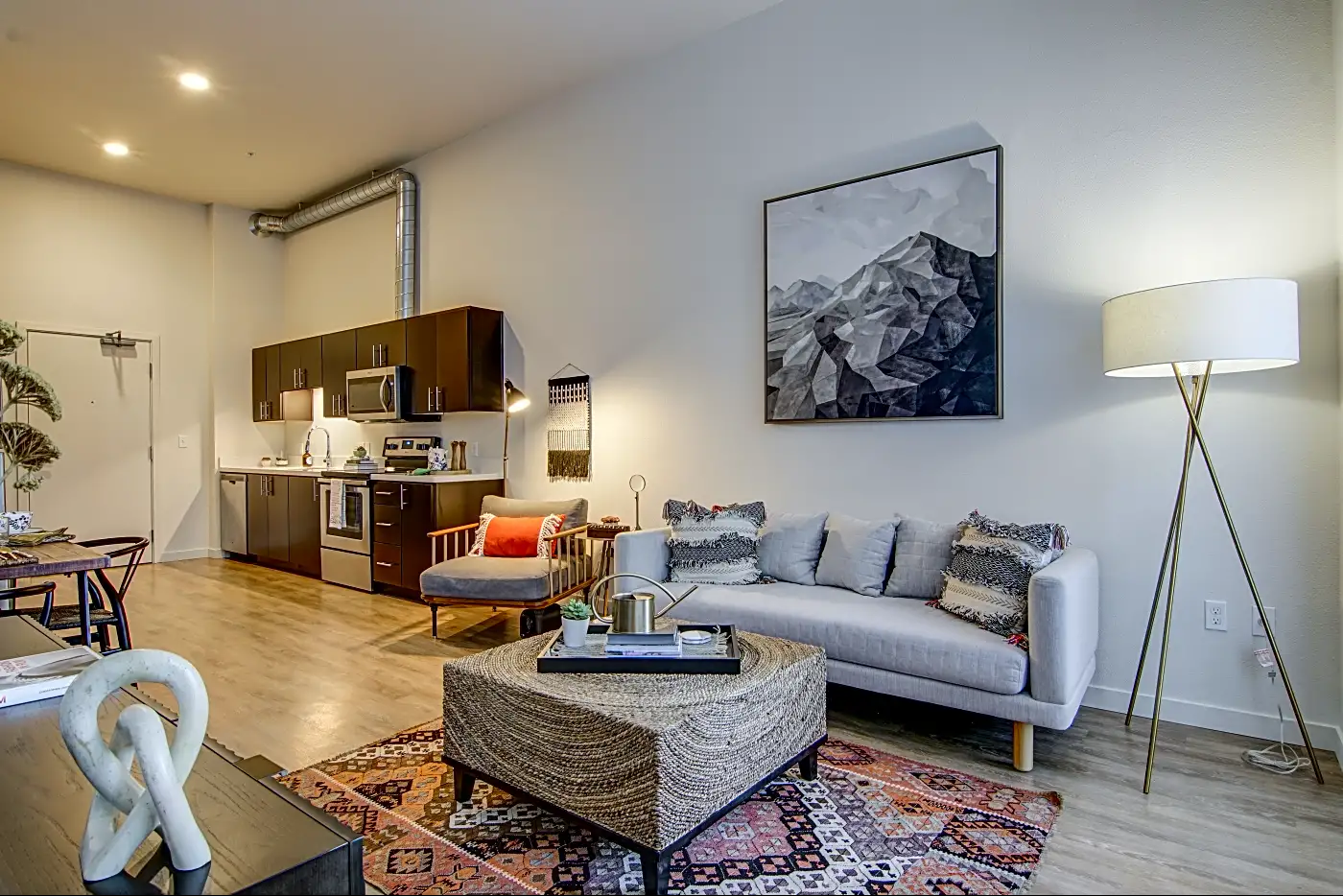 A modern living room and kitchenette area with a beige couch, patterned pillows, a round textured coffee table, a large artwork of mountains on the wall, a tall white floor lamp, and a dark wood kitchen with stainless steel appliances and cabinets