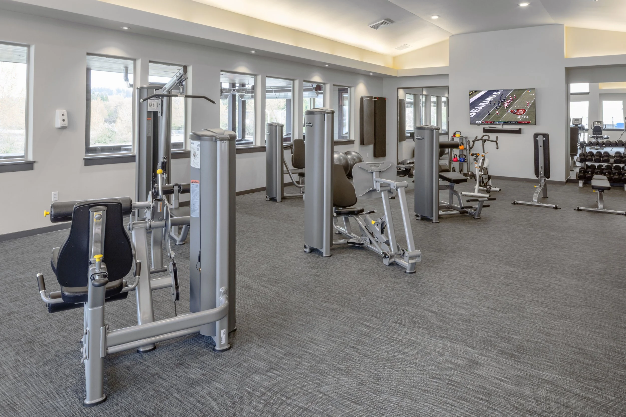 Empty gym with exercise equipment, weight machines, free weights, and a television on the wall.