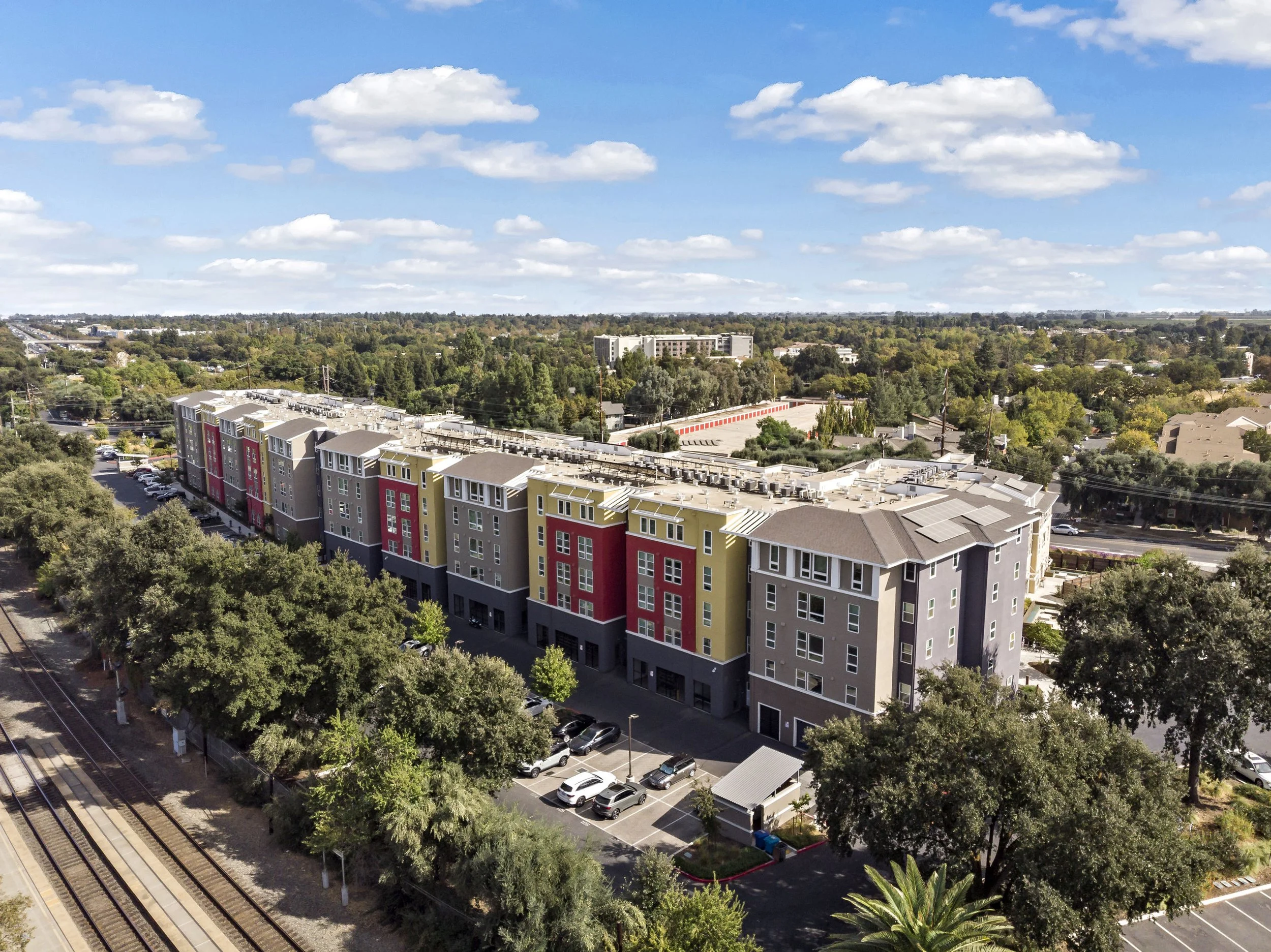 Colorful modern apartment building with trees and parking lot, set in suburban area with train tracks nearby and cloudy sky.