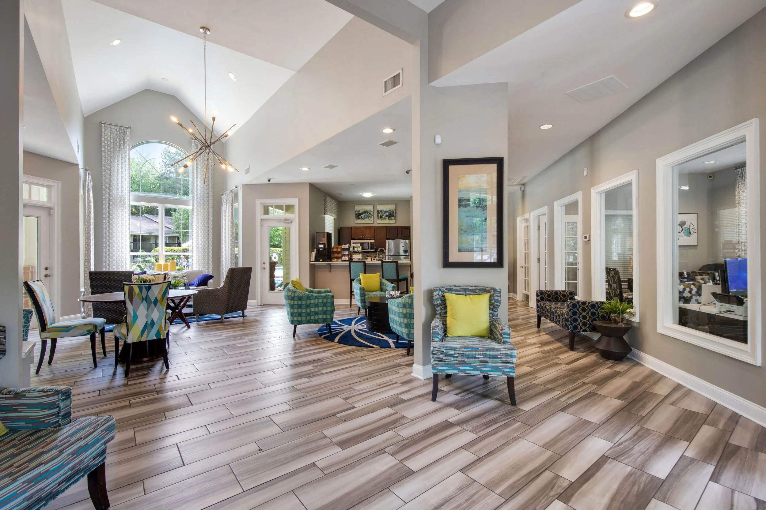 Spacious modern living area with high vaulted ceiling, large windows, and wooden flooring. Features various chairs including a patterned armchair with a yellow pillow, a blue plaid armchair, and a set of dining chairs. There is a circular rug, bar se