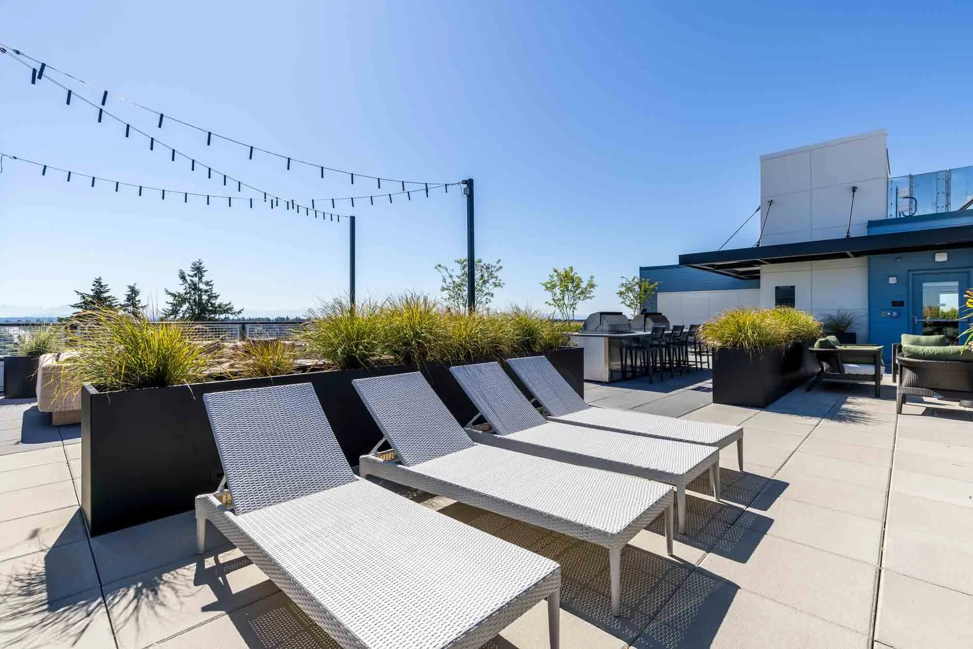 Rooftop patio with lounge chairs, potted plants, string lights, outdoor dining table, grill, and seating area under a clear blue sky.